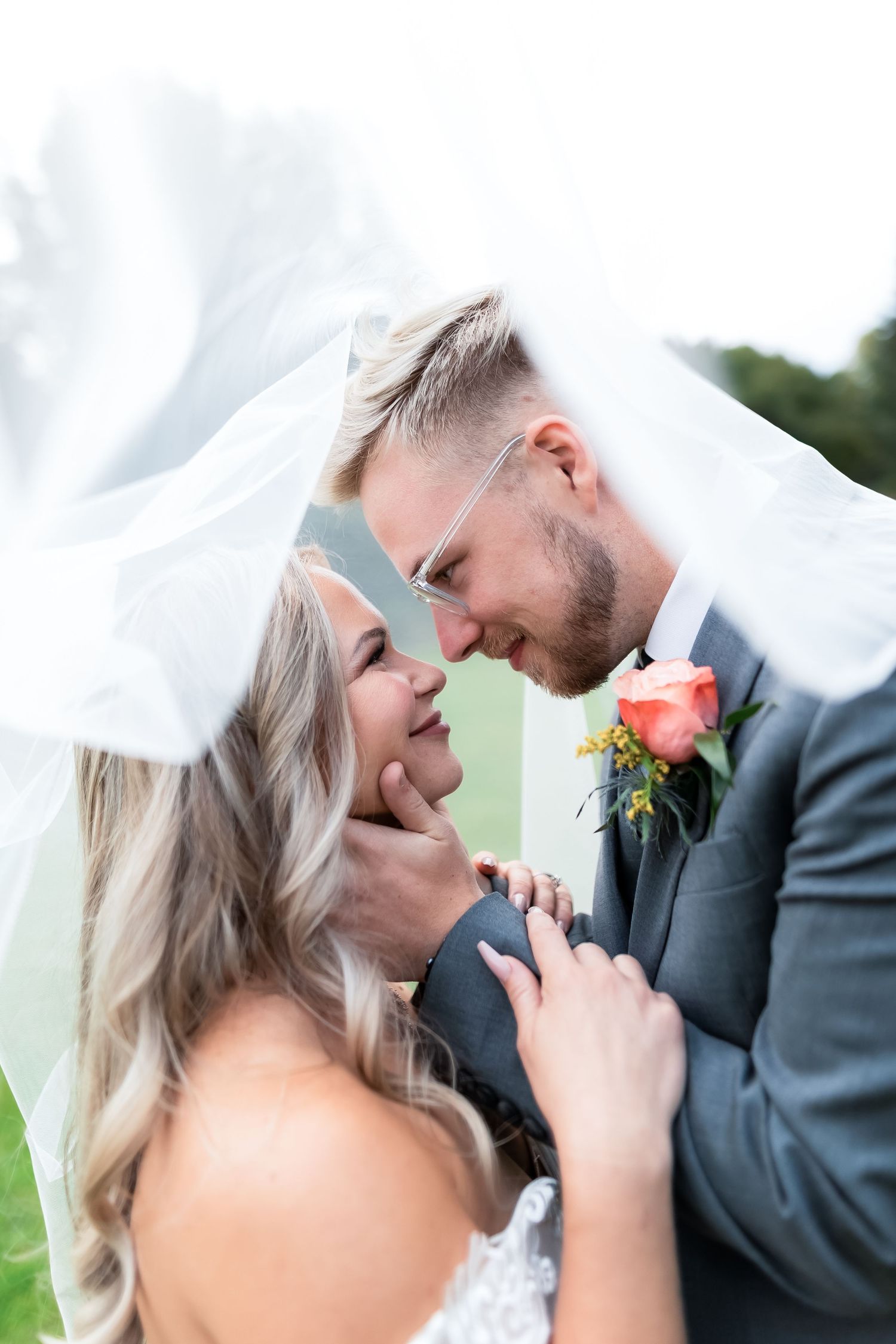 Bride and groom share a romantic moment under a flowing veil, with the groom wearing a gray suit and coral boutonniere.