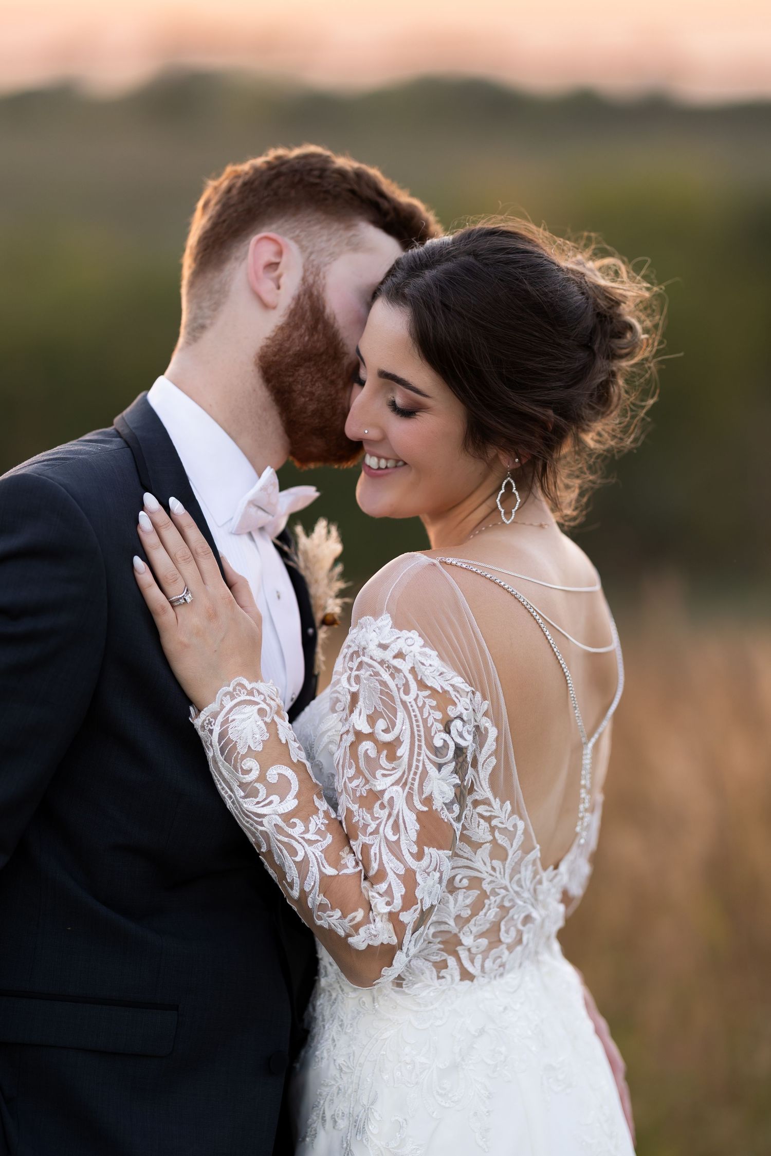 A romantic wedding couple embraces in an outdoor field during sunset while wearing formal attire.