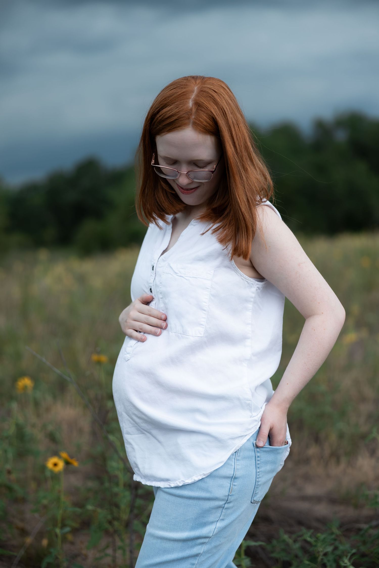 Pregnant redhead woman in white sleeveless shirt and jeans standing in a field, gently holding her baby bump.
