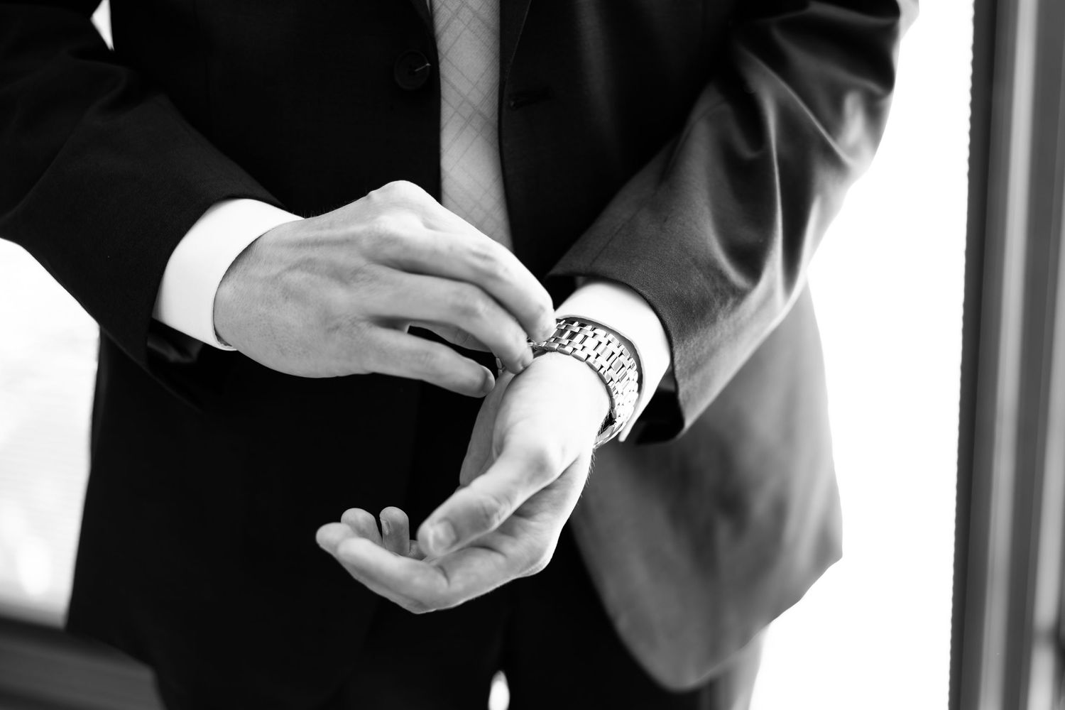 Man in black suit adjusting cufflinks on his wrist, wearing a watch, black and white photo.