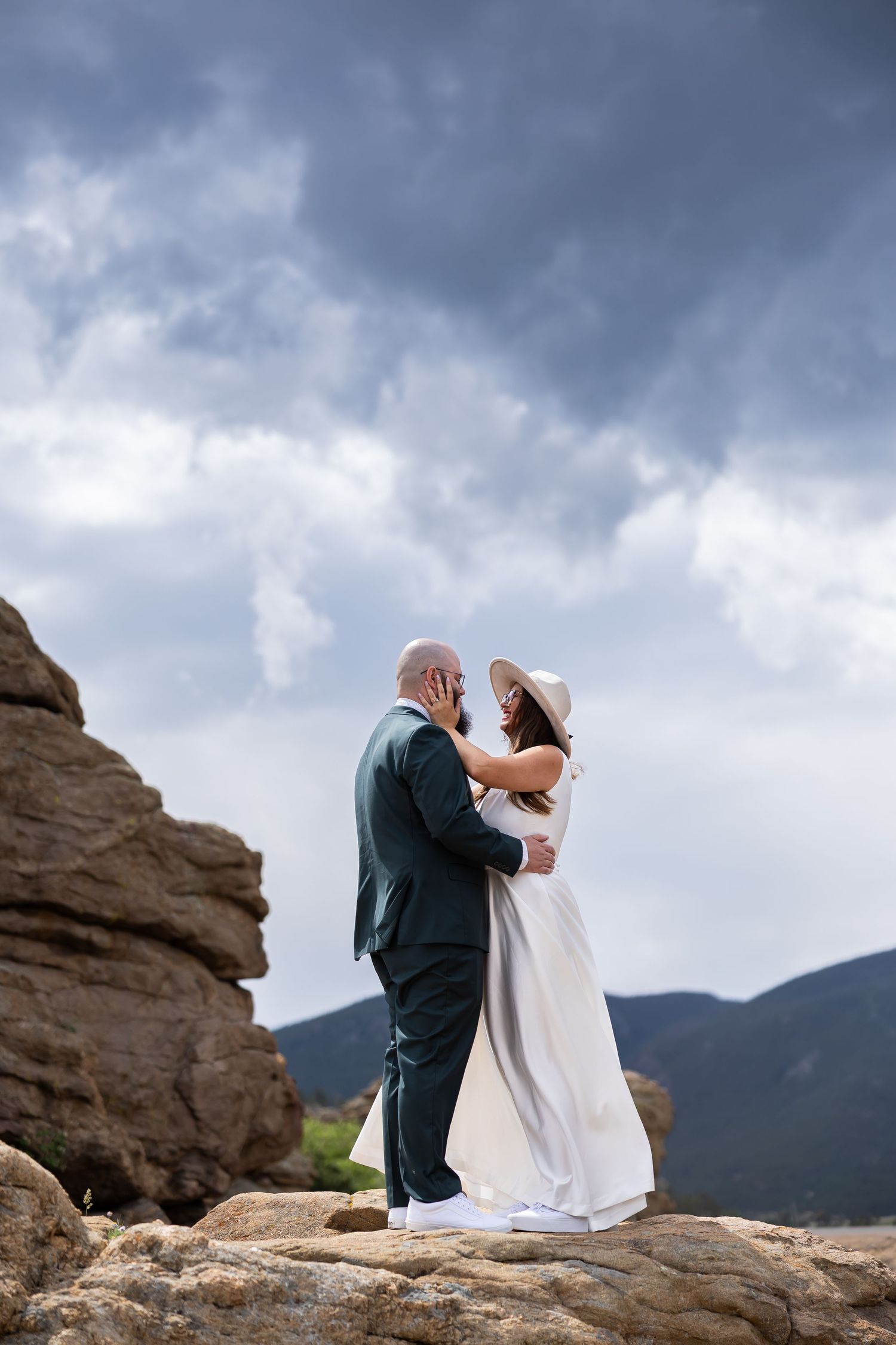 Wedding couple stands on dramatic rocky overlook against stormy mountain backdrop.