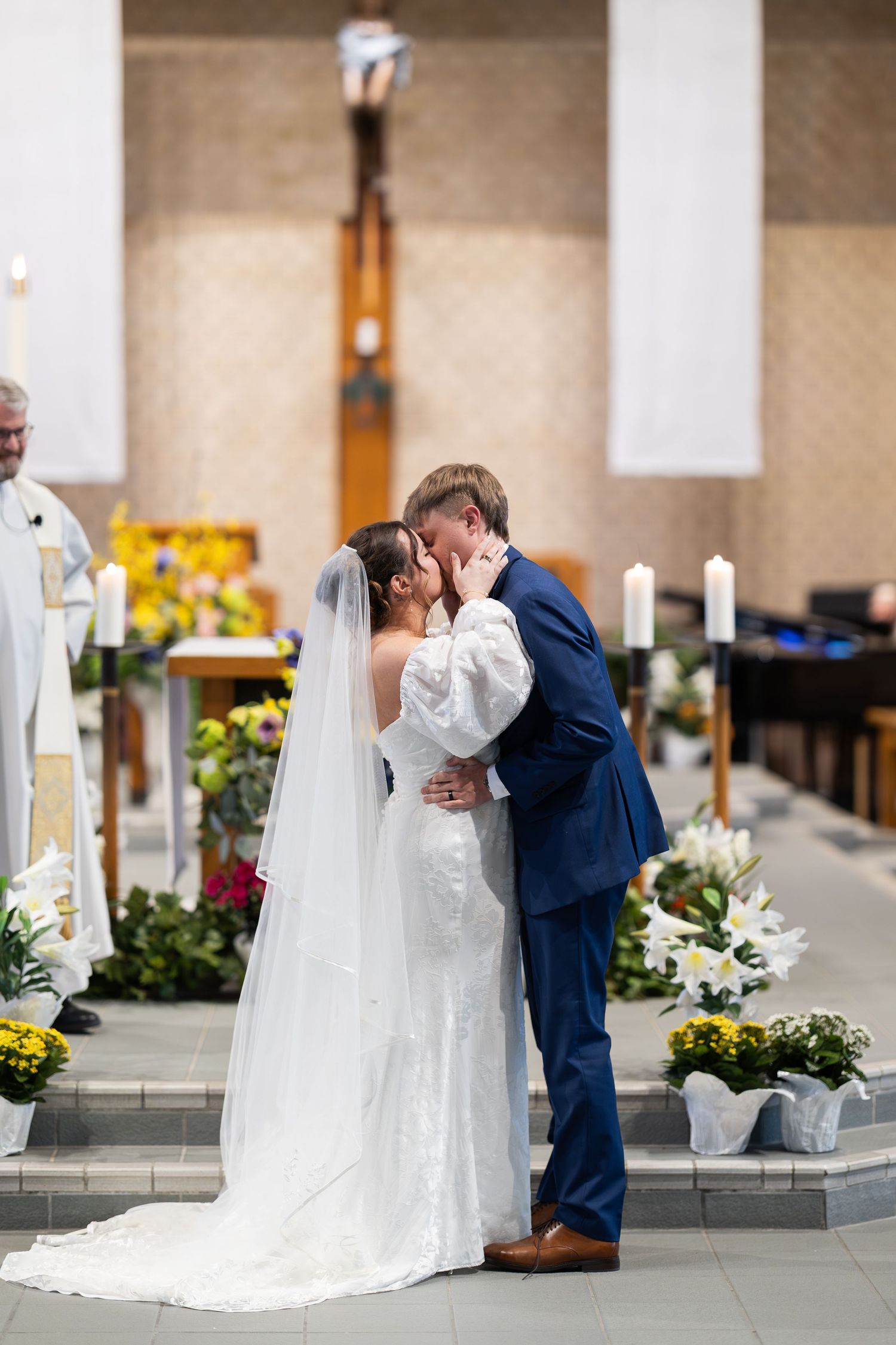 Bride and groom share their first kiss at the altar during a church wedding ceremony, surrounded by floral arrangements.