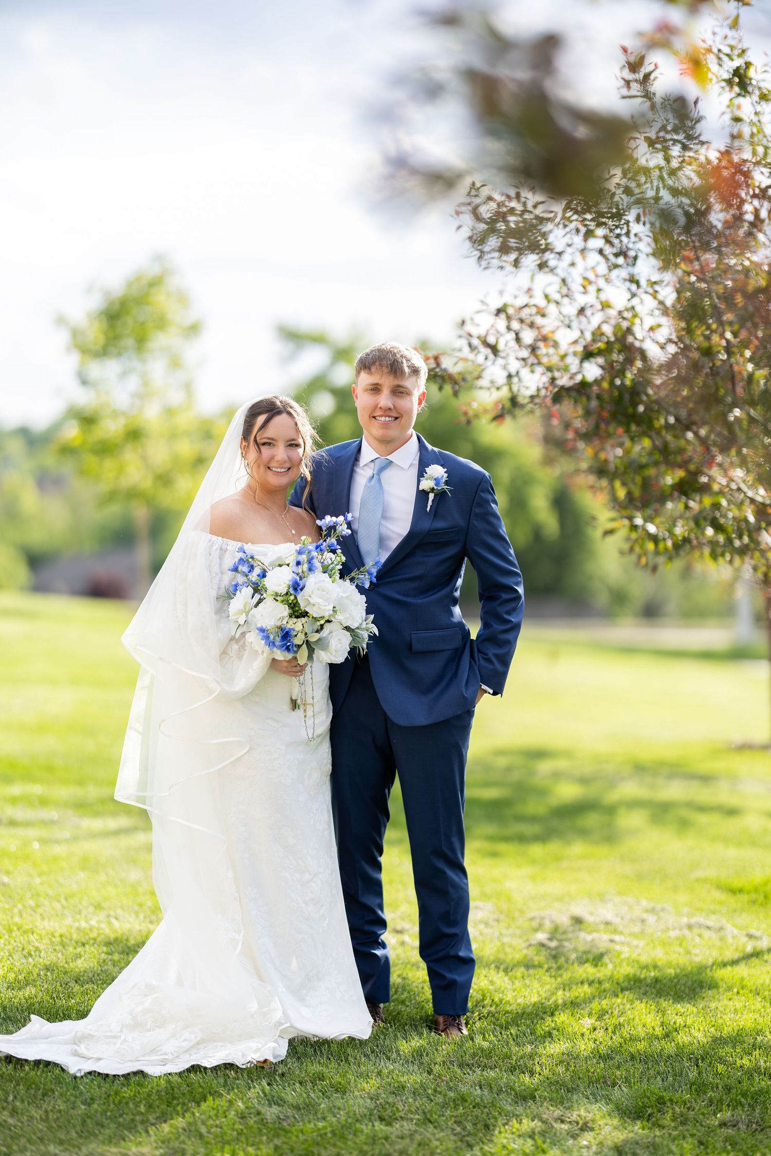 Bride and groom posing outdoors on green grass, bride holding blue and white bouquet, groom in navy suit.