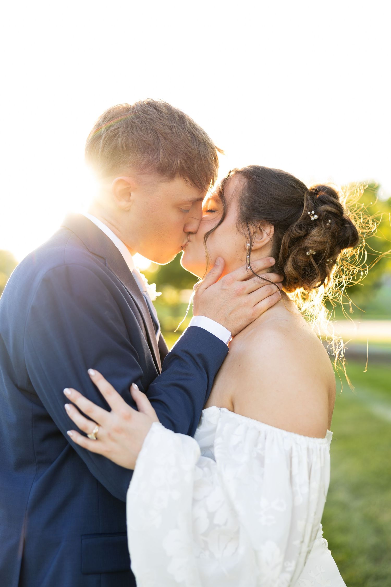 A bride and groom share a romantic kiss outdoors, bathed in golden sunlight, with the groom in a navy suit and bride in a white gown.