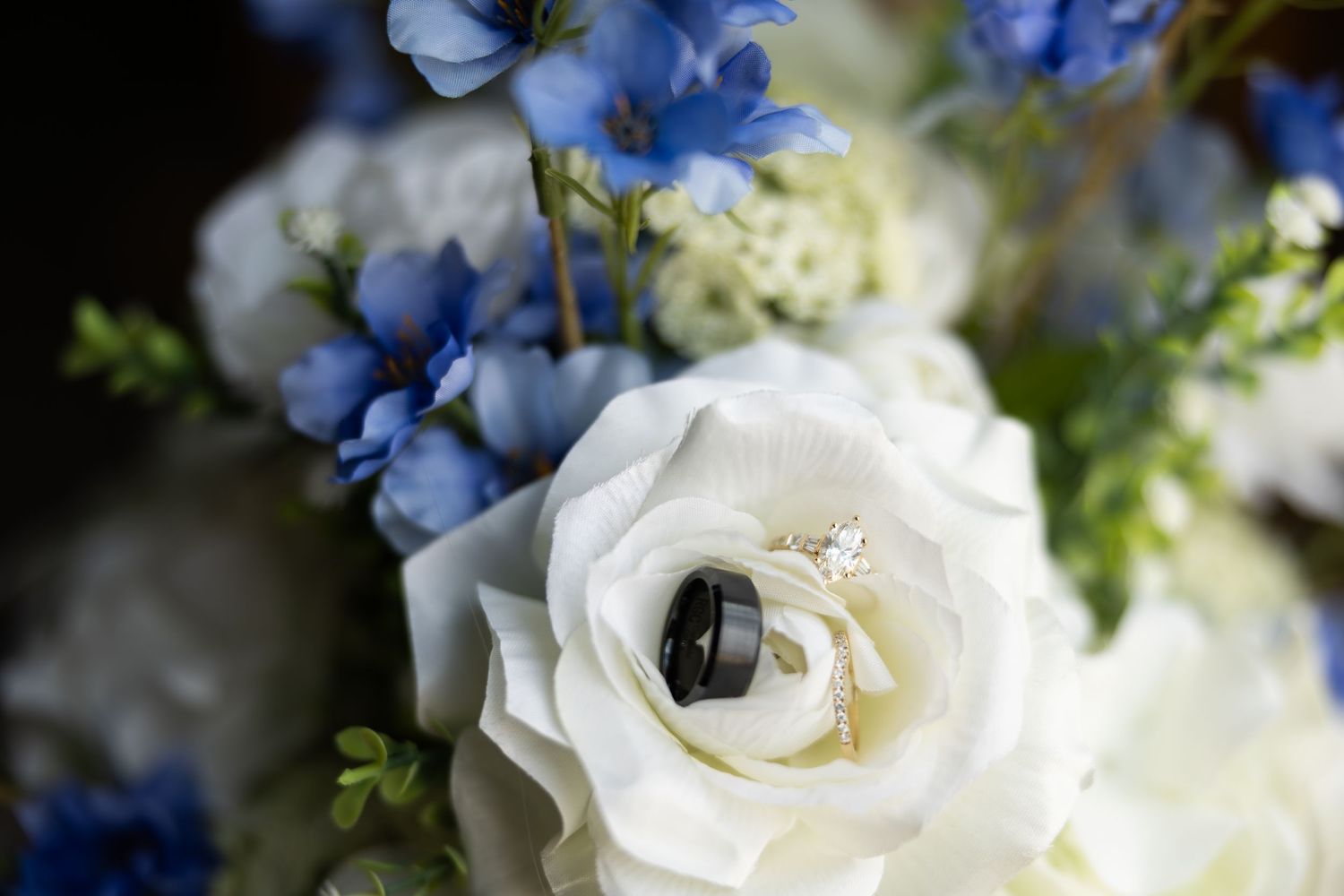 Wedding rings nestled in a white rose surrounded by blue delphinium flowers in a bridal bouquet.