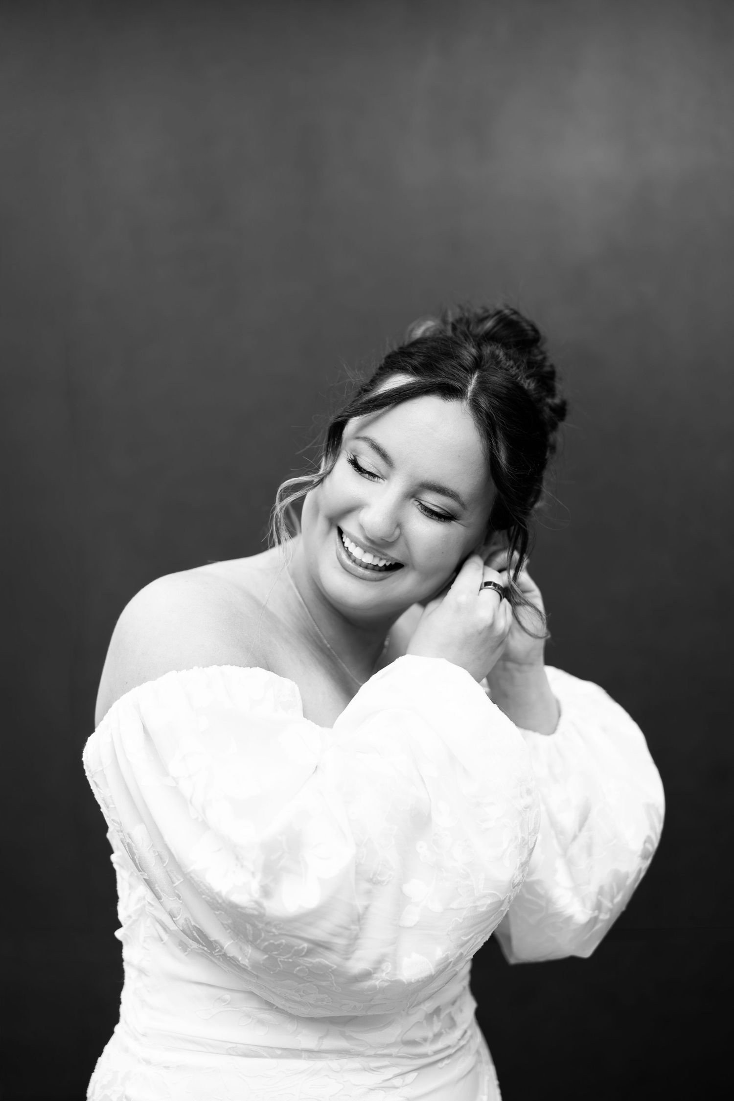 Black and white portrait of a smiling bride in an off-shoulder white gown, touching her earring.