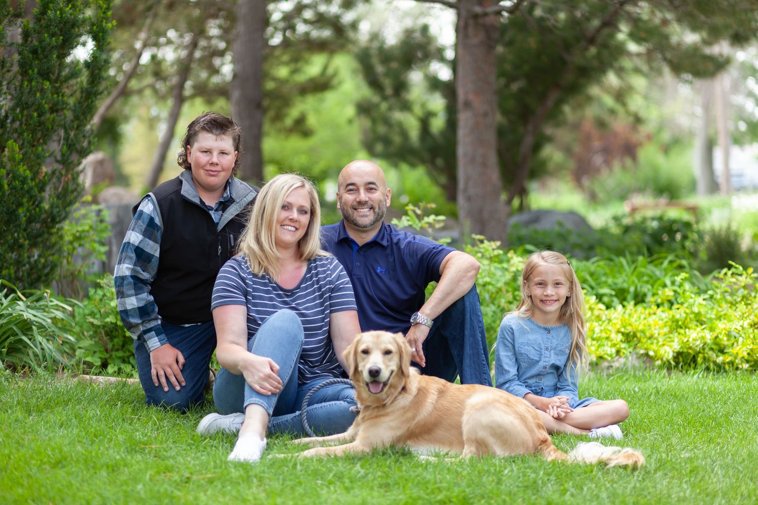Happy family sitting in lush garden with golden retriever dog on sunny day.