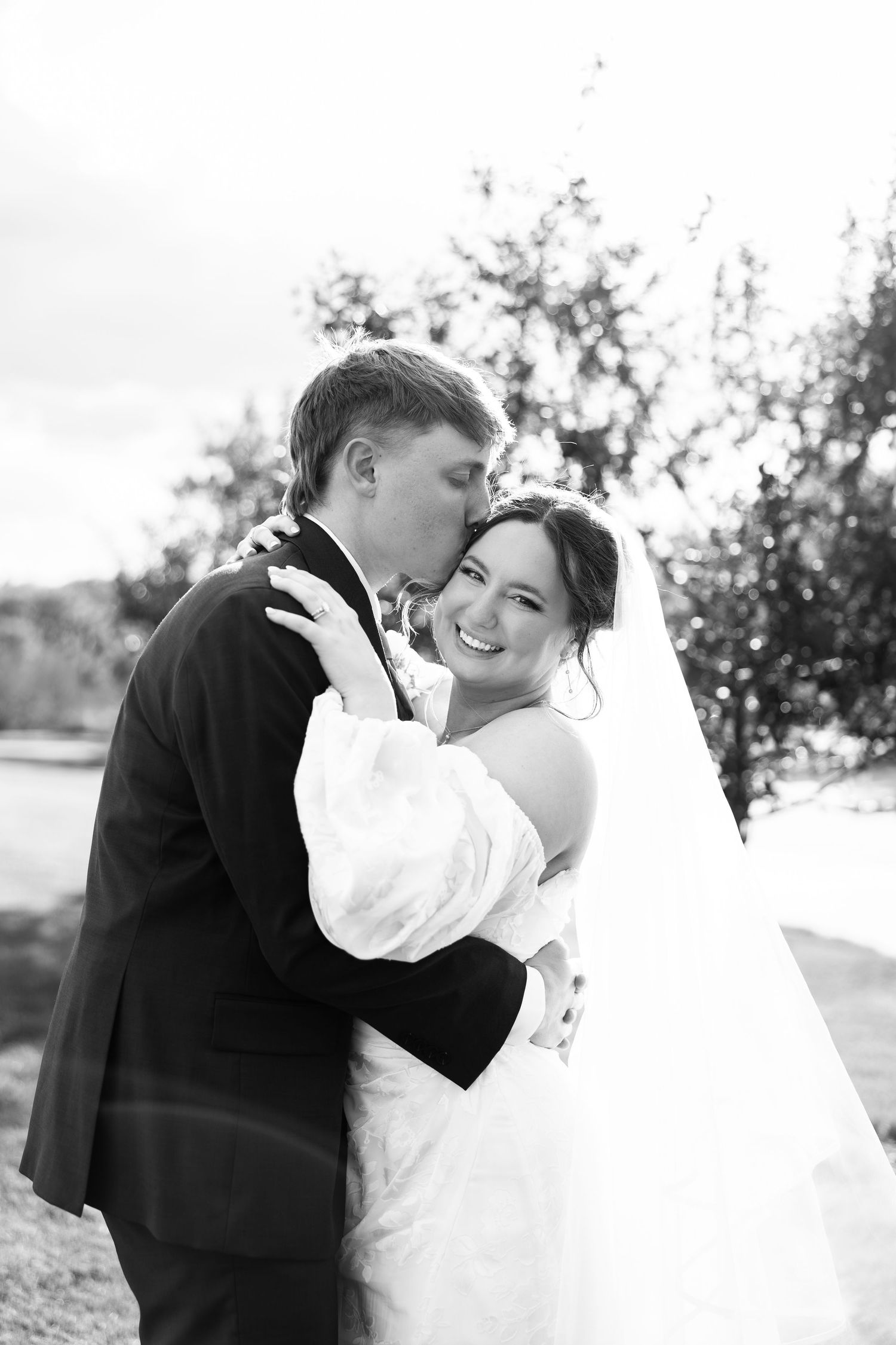 Black and white photo of a groom kissing his bride on the cheek as she smiles joyfully on their wedding day.