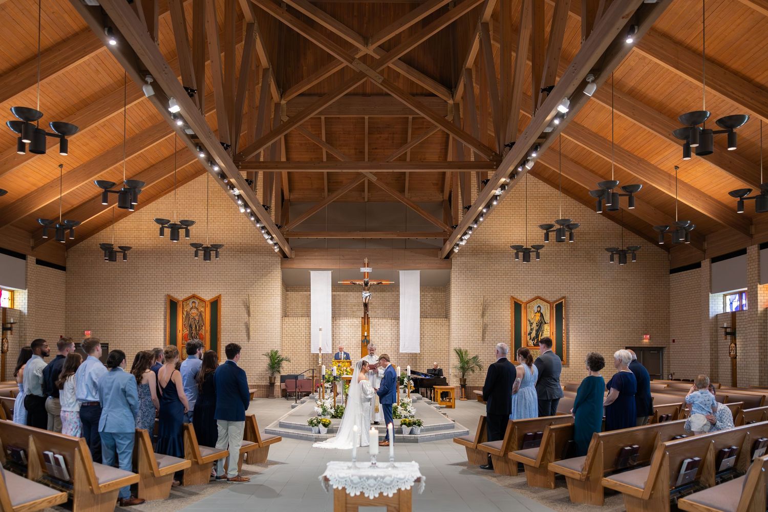 Elegant church wedding ceremony with guests standing in wooden pew-lined aisle as couple exchanges vows at floral altar.