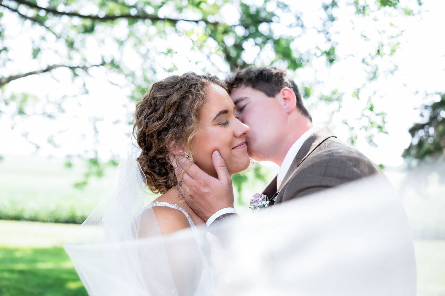 A groom tenderly cups his bride's face for a kiss, her veil flowing in the breeze at an outdoor wedding.
