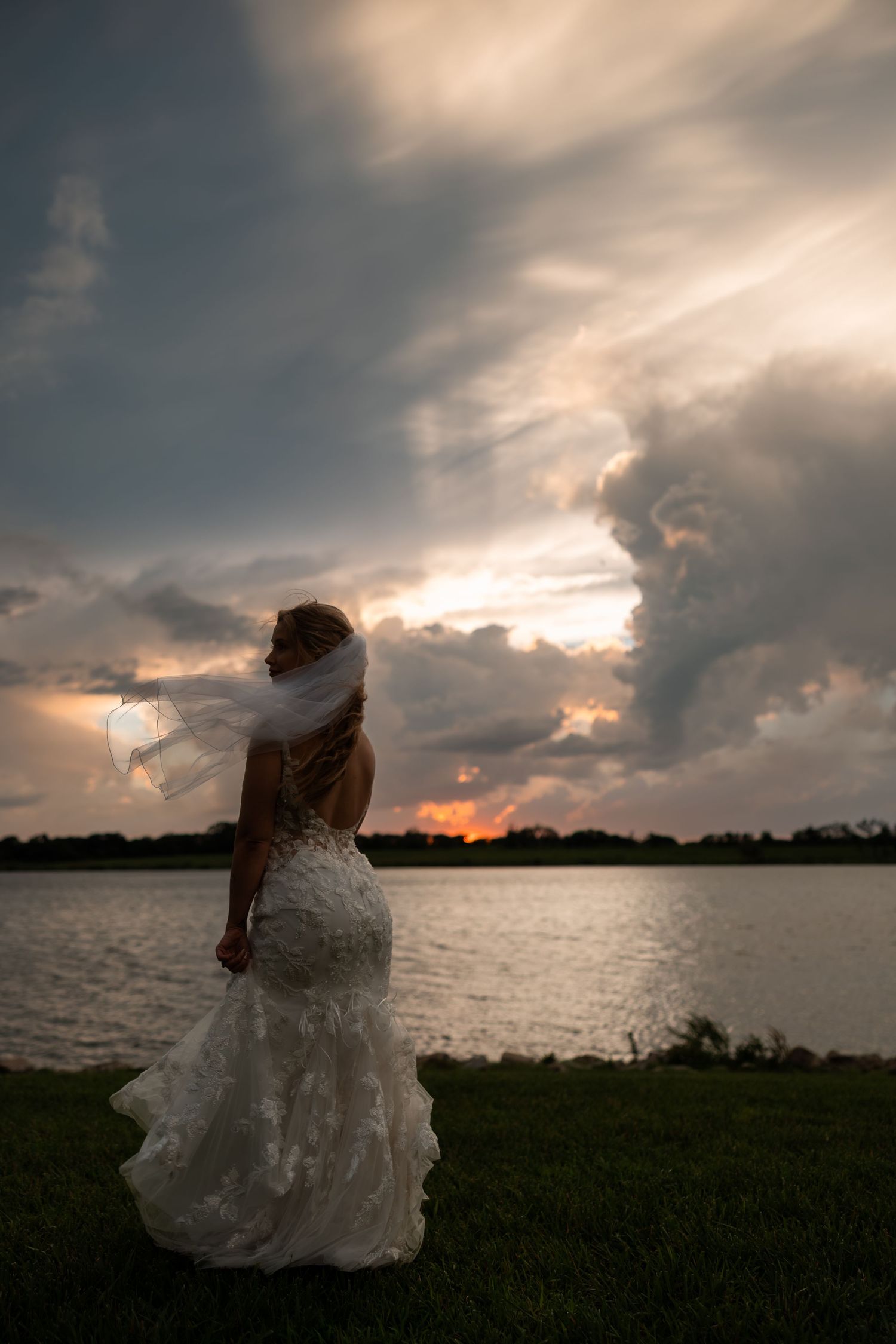 Bride in white lace gown and veil stands by a lake at sunset under dramatic stormy clouds.