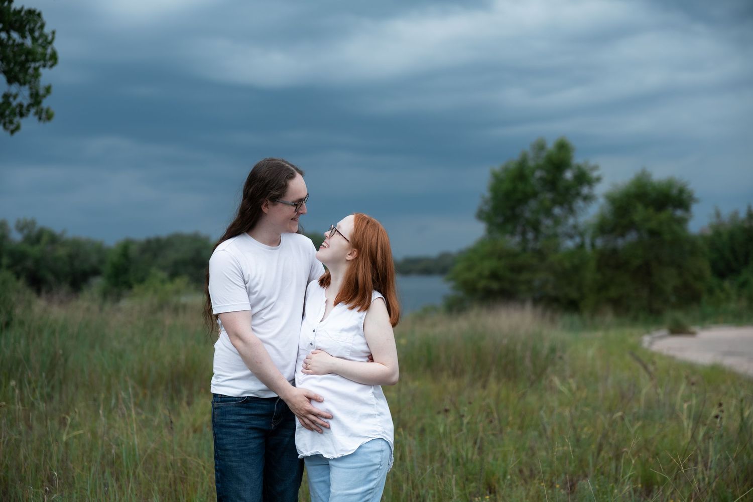 A couple in matching white shirts embraces in a green field under dramatic cloudy skies, the woman appearing pregnant.