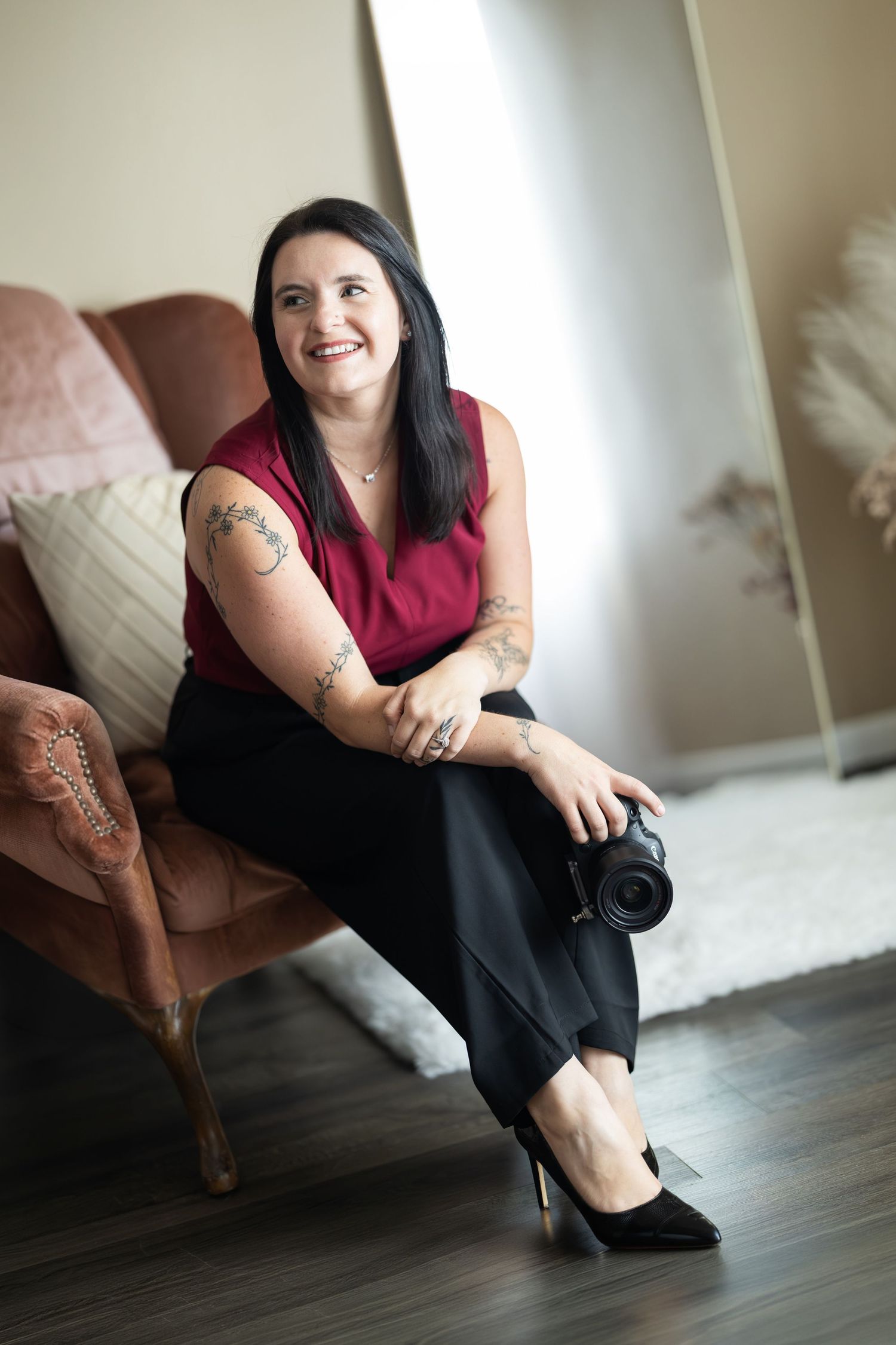A smiling woman with tattoos sits in a chair holding a camera, wearing a red top and black pants with heels.