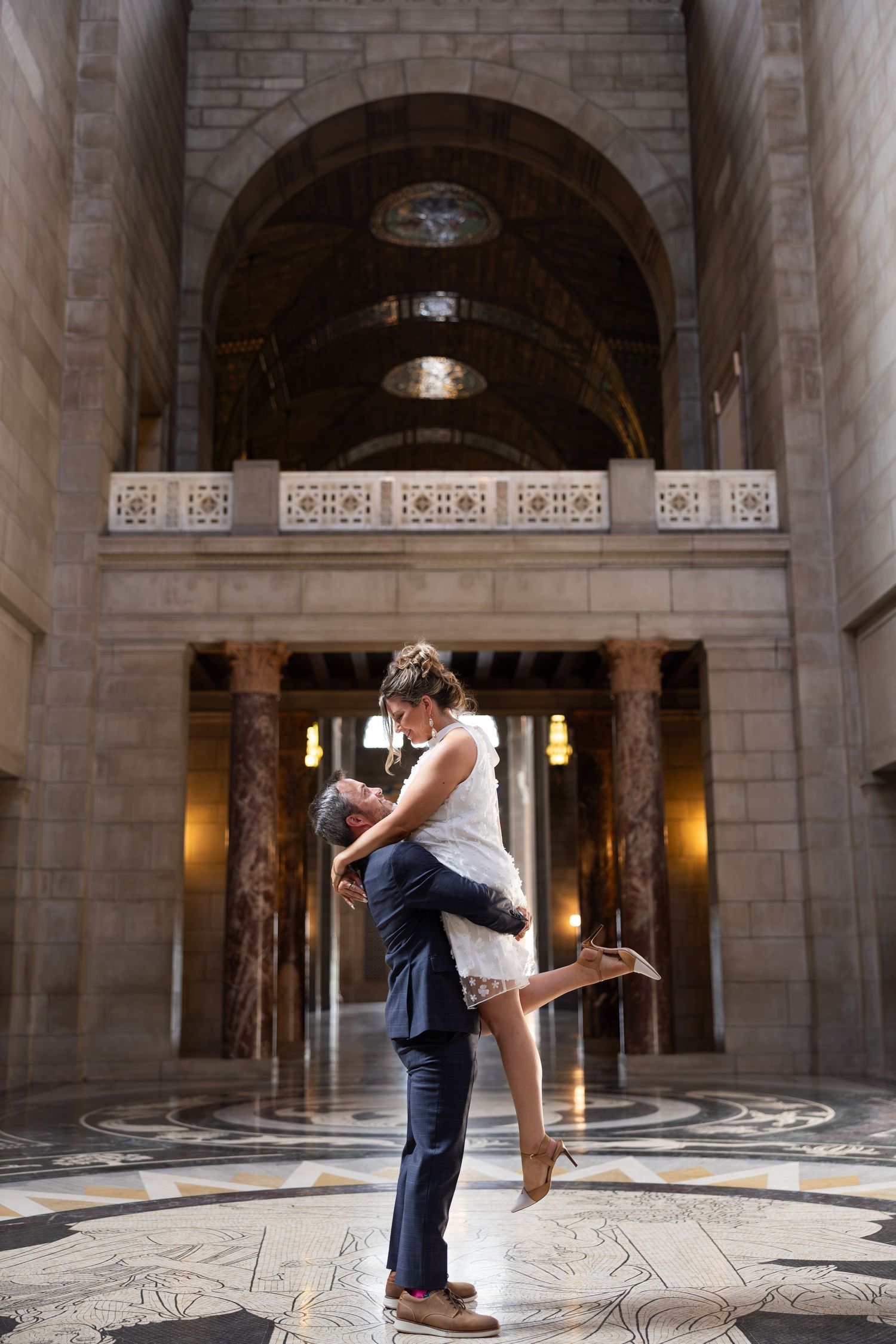 Groom lifts bride in elegant marble hall with arched ceilings during romantic wedding photo session.