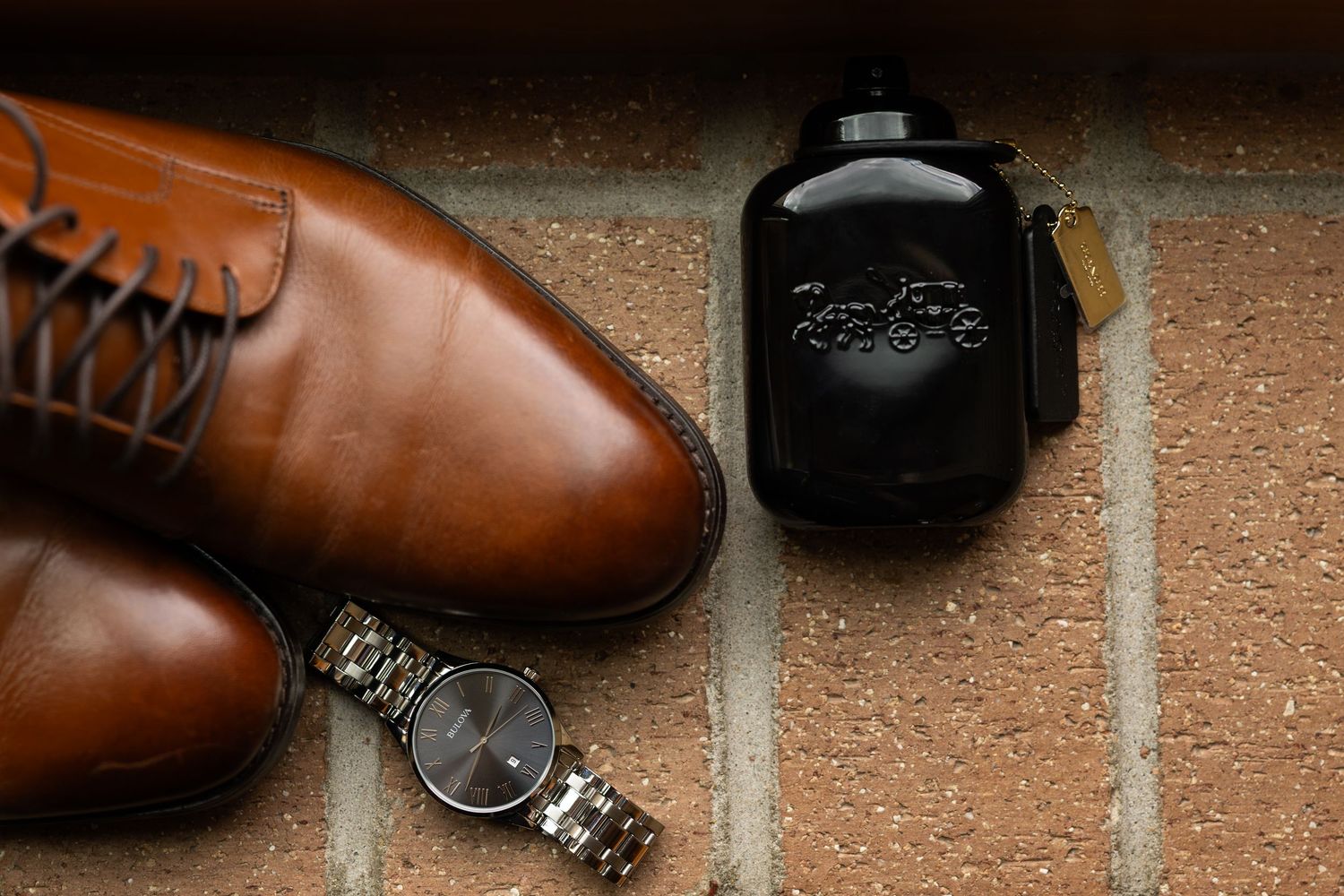 Brown leather dress shoes, silver watch, and black flask arranged on brick surface.