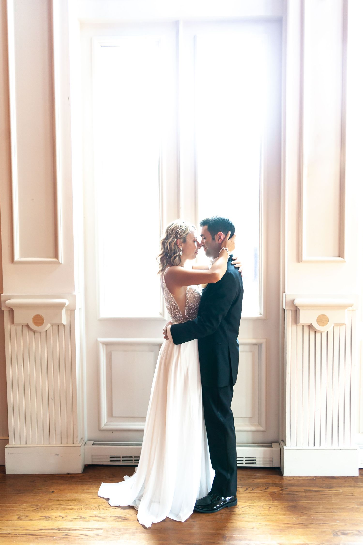 Elegant couple embracing in sunlit room with classic white wedding attire and hardwood floors.