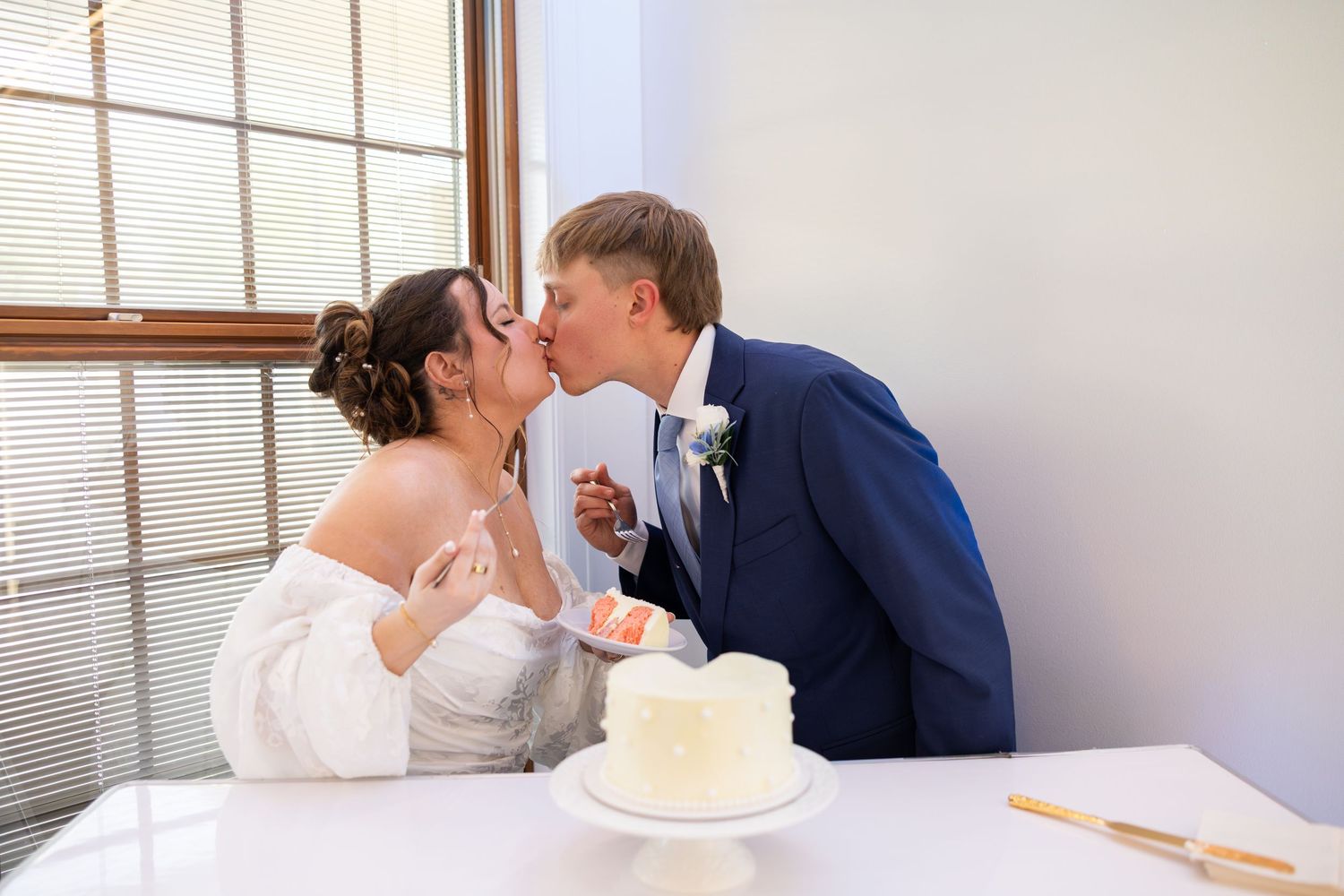 Bride and groom sharing a kiss at wedding cake cutting table, white cake in foreground, bright window background.