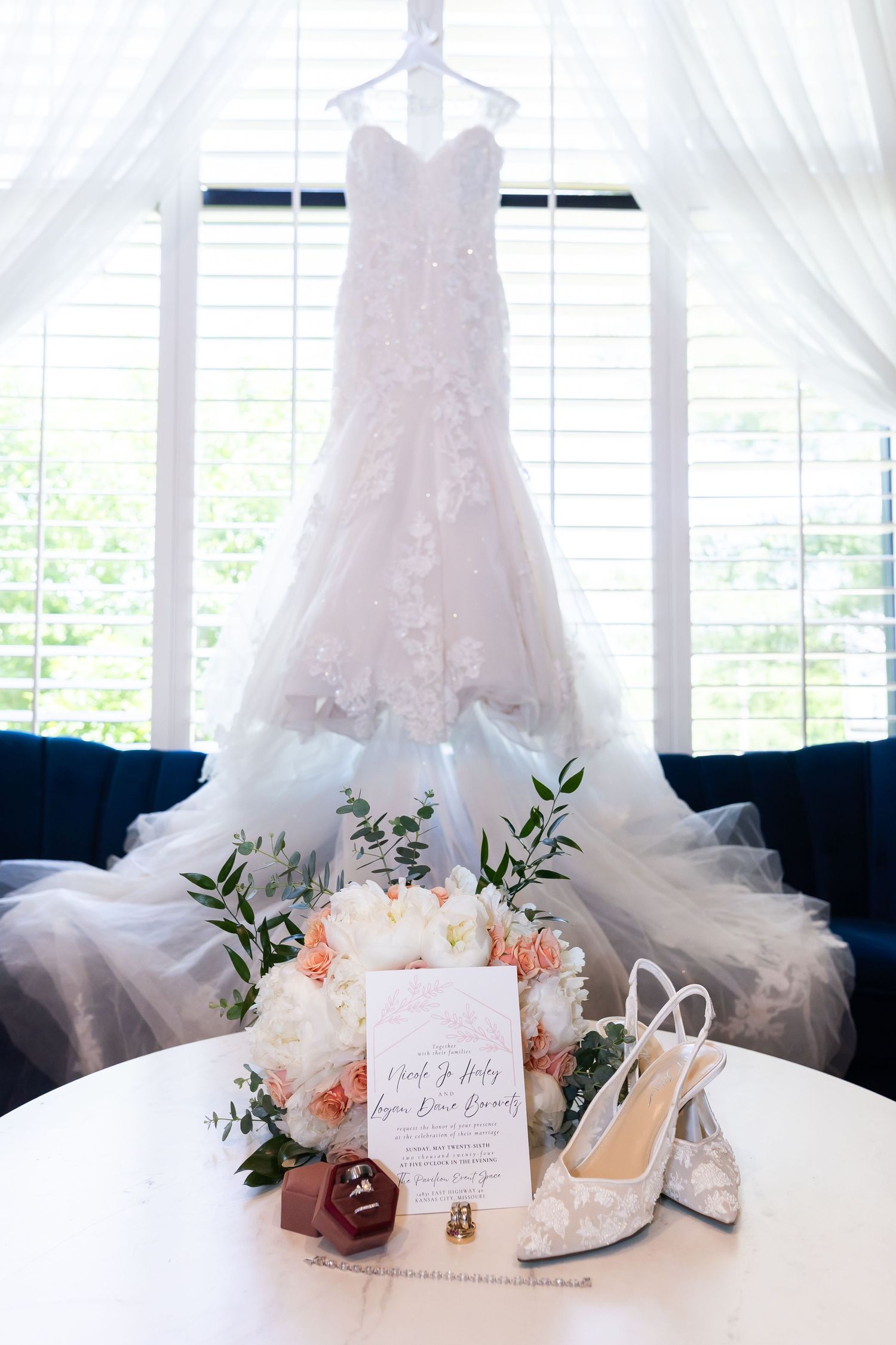 Wedding dress hanging by window with bridal accessories and floral arrangement below.
