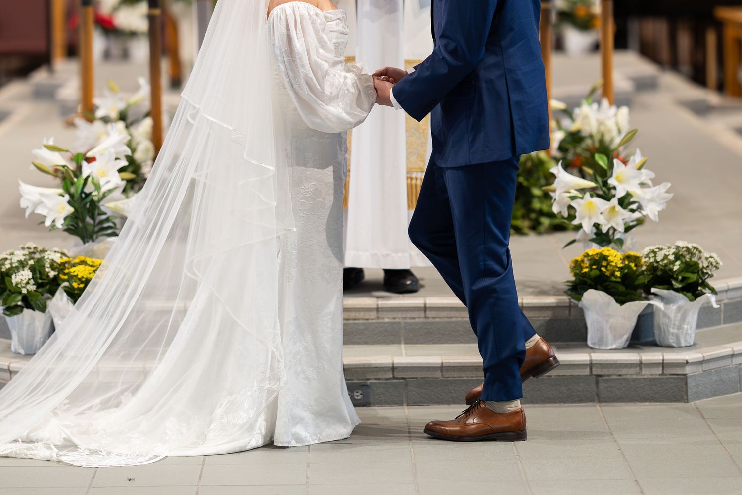 Bride in white gown and groom in blue suit holding hands at floral church altar during wedding ceremony.