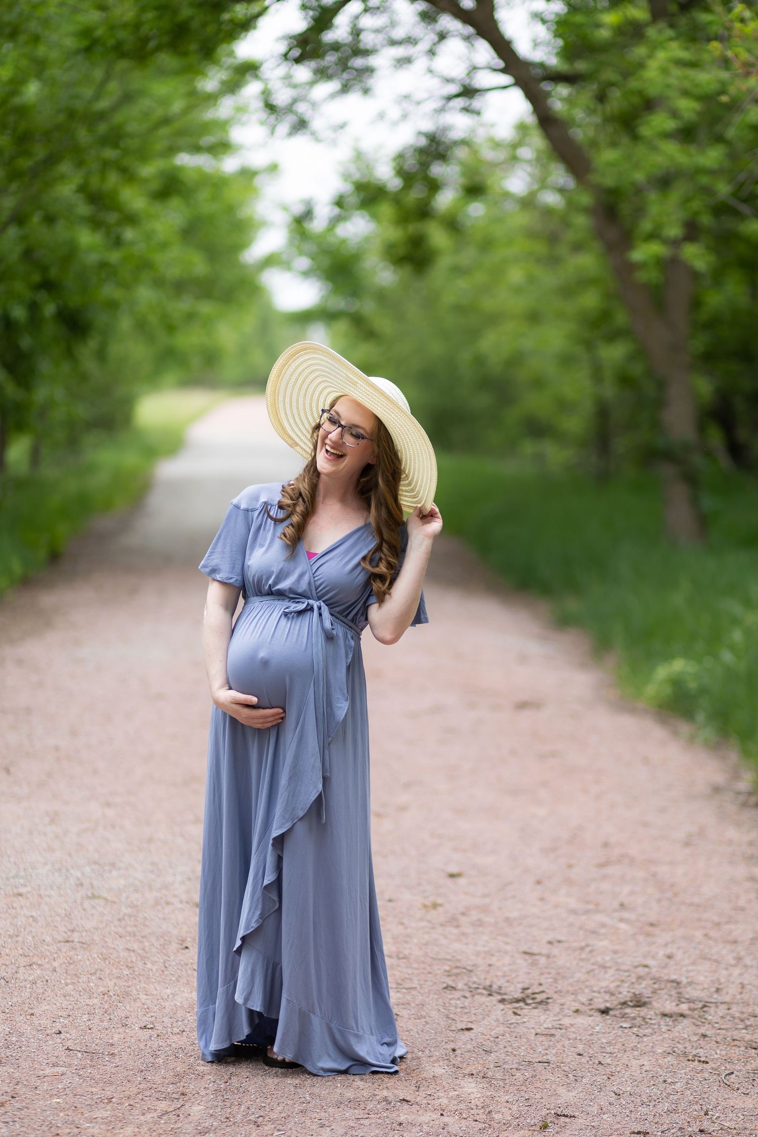 A maternity photo of a woman in flowing blue dress posing on a tree-lined path wearing a sun hat.