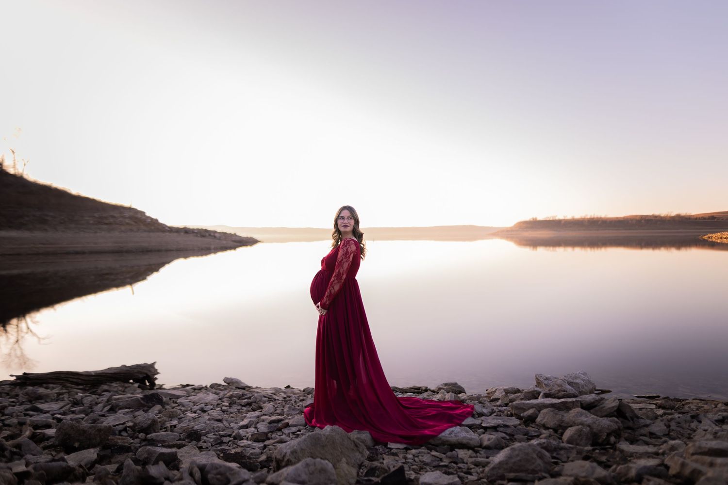 A maternity photo of a woman in a burgundy dress standing by a lake during golden hour.