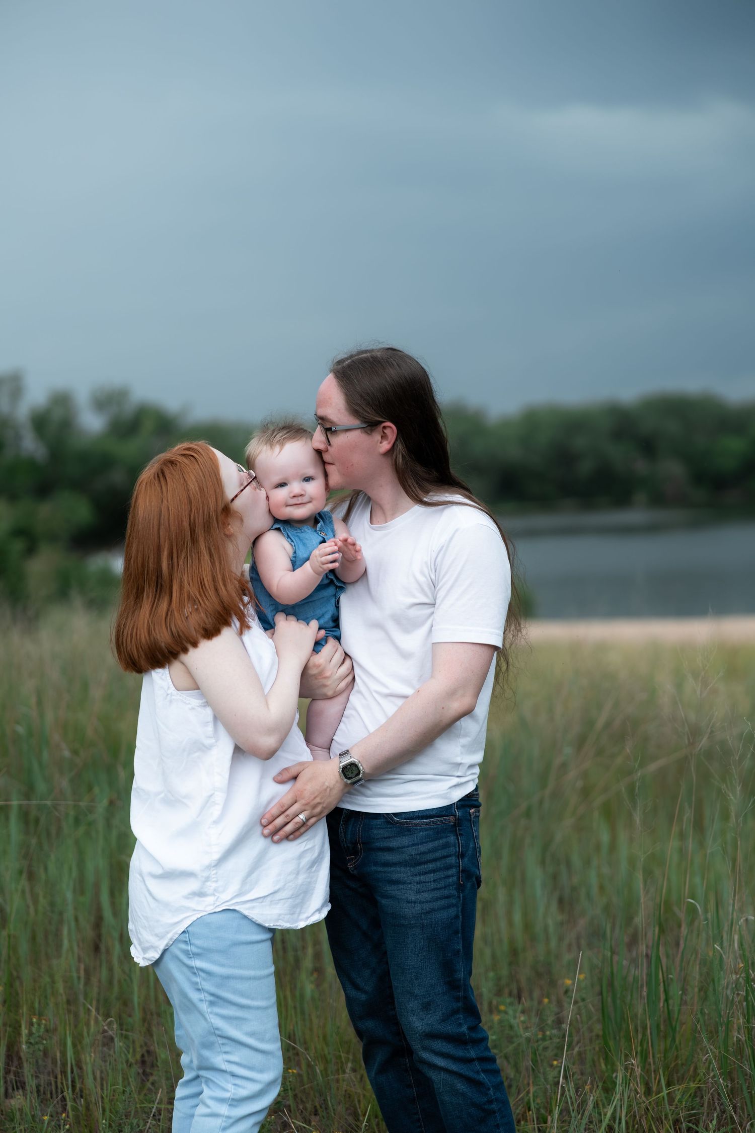 A couple in white shirts kiss while holding their smiling baby outdoors near a lake with stormy skies.