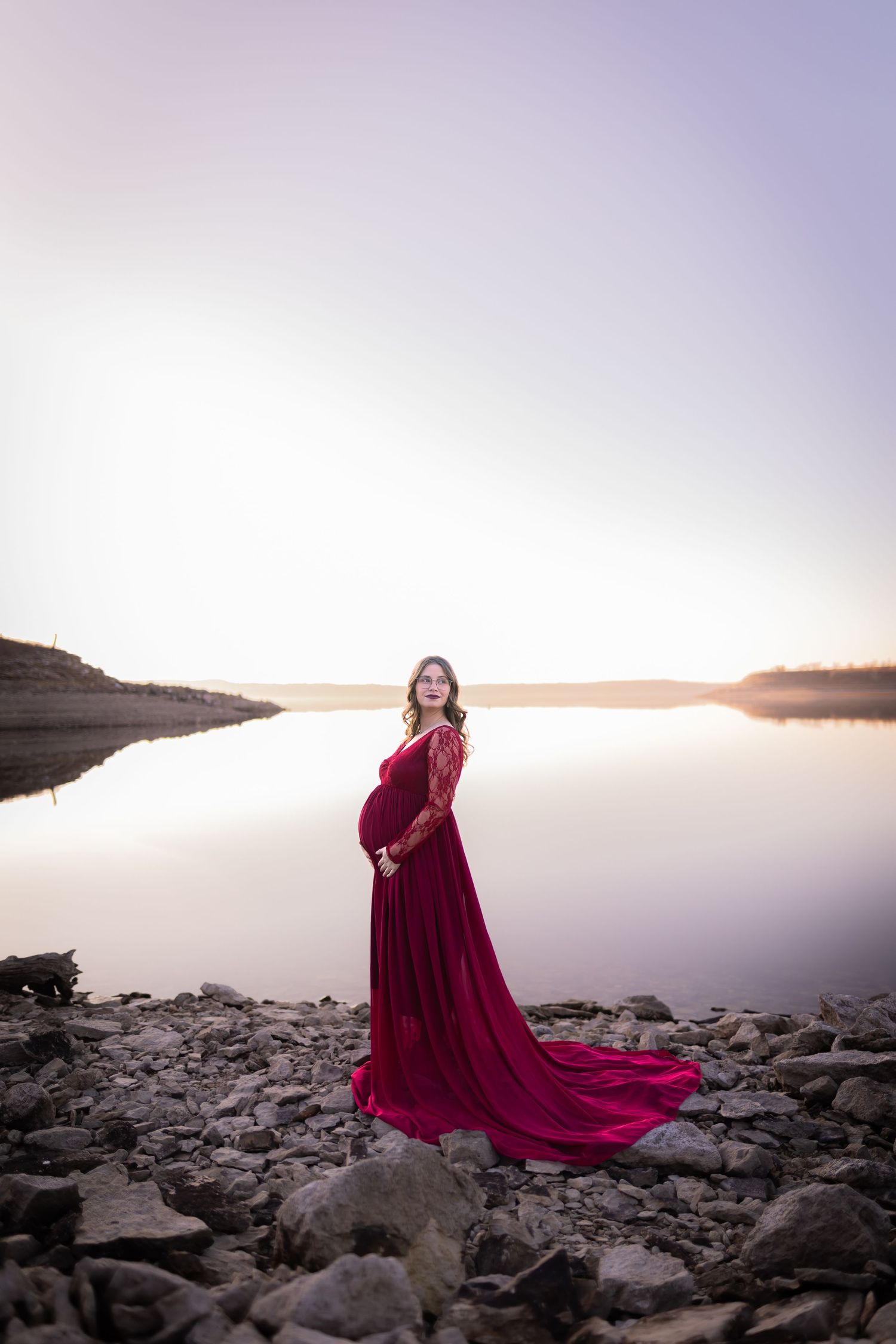 Silhouette of maternity photo in flowing burgundy dress against serene lake at sunset.
