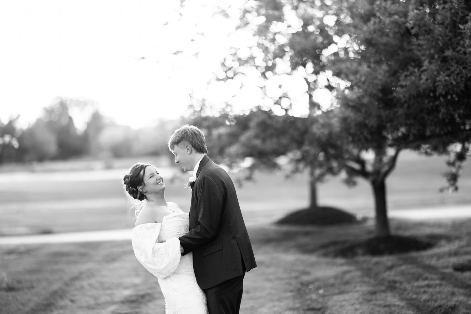 Black and white photo of a bride and groom sharing a romantic moment outdoors near a tree on their wedding day.