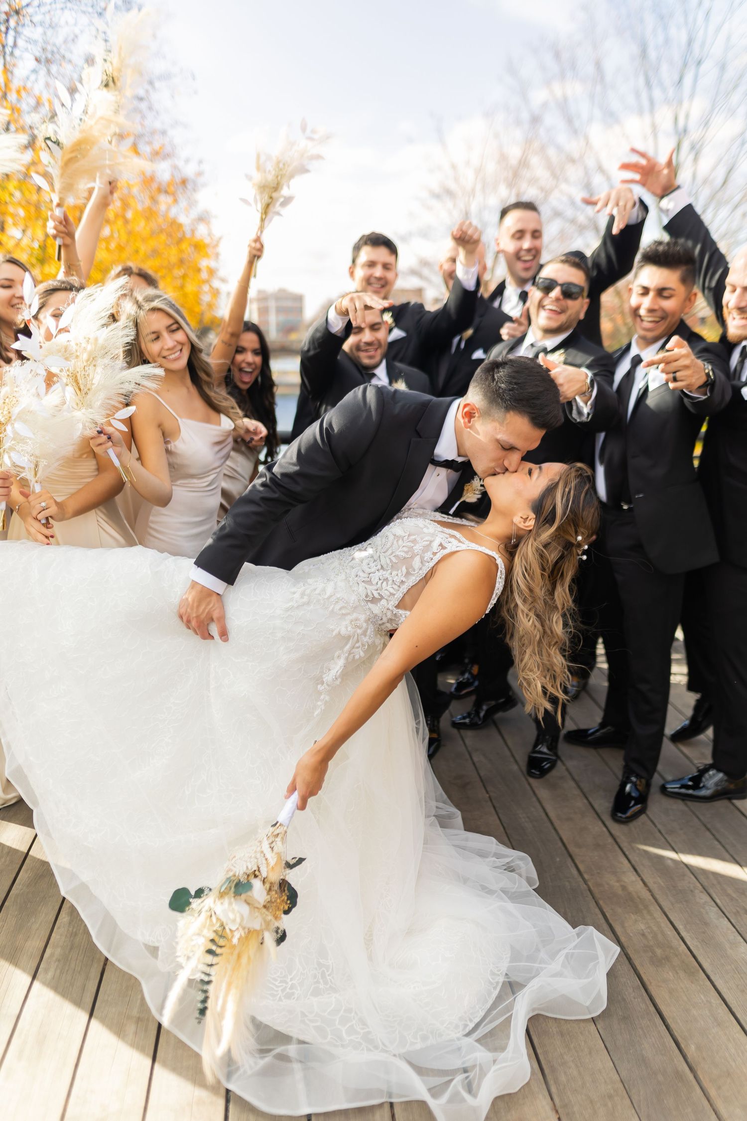A wedding party celebrates joyfully on a wooden deck during autumn with the newlyweds sharing a romantic dip.
