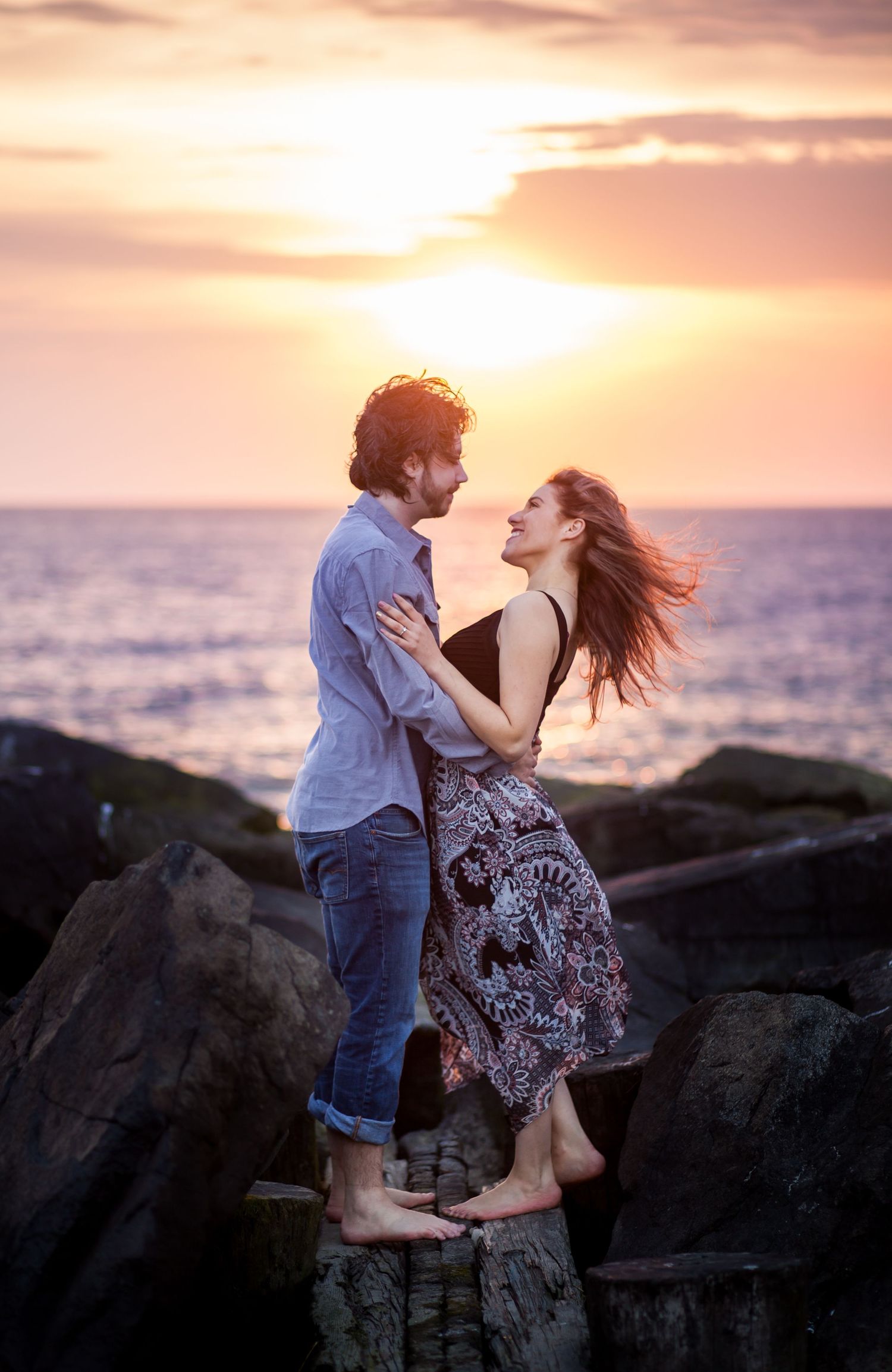 A romantic couple embracing on coastal rocks during a vibrant sunset over the ocean.