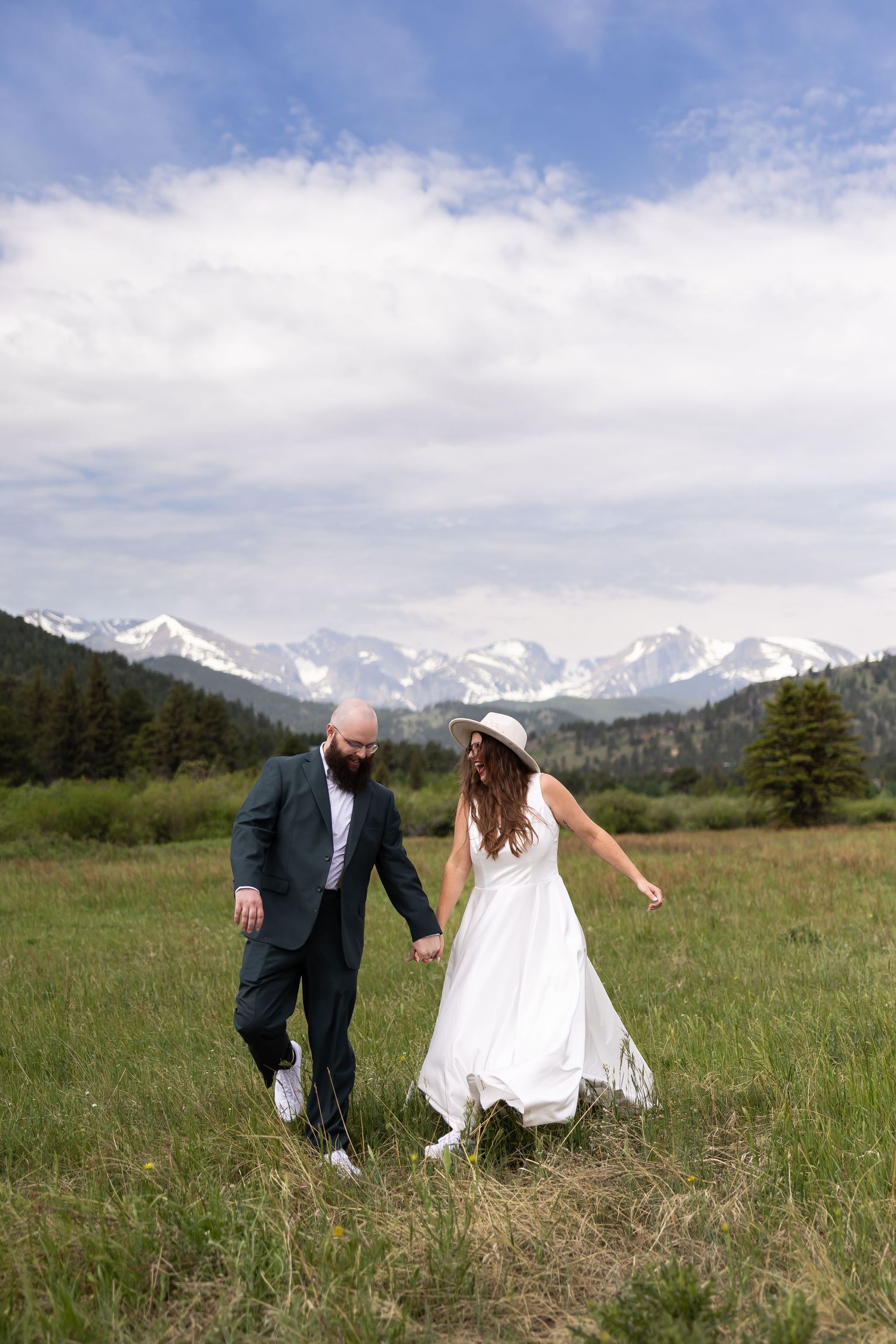 Newlyweds running playfully through a meadow with snow-capped mountains in the background.