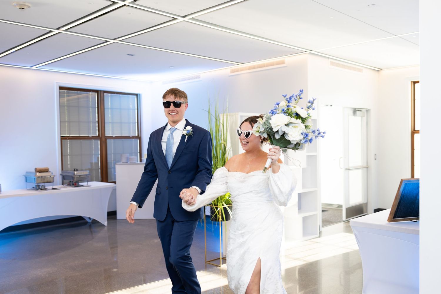 Bride and groom walk hand-in-hand wearing sunglasses, bride holding blue and white bouquet at indoor wedding reception.