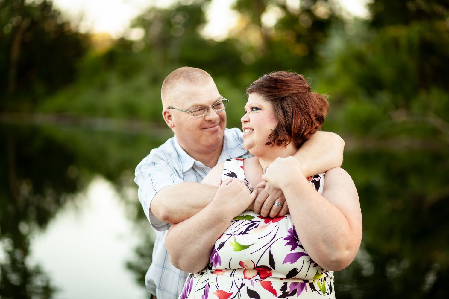 Couple shares a joyful embrace while laughing together by a tranquil pond at sunset.