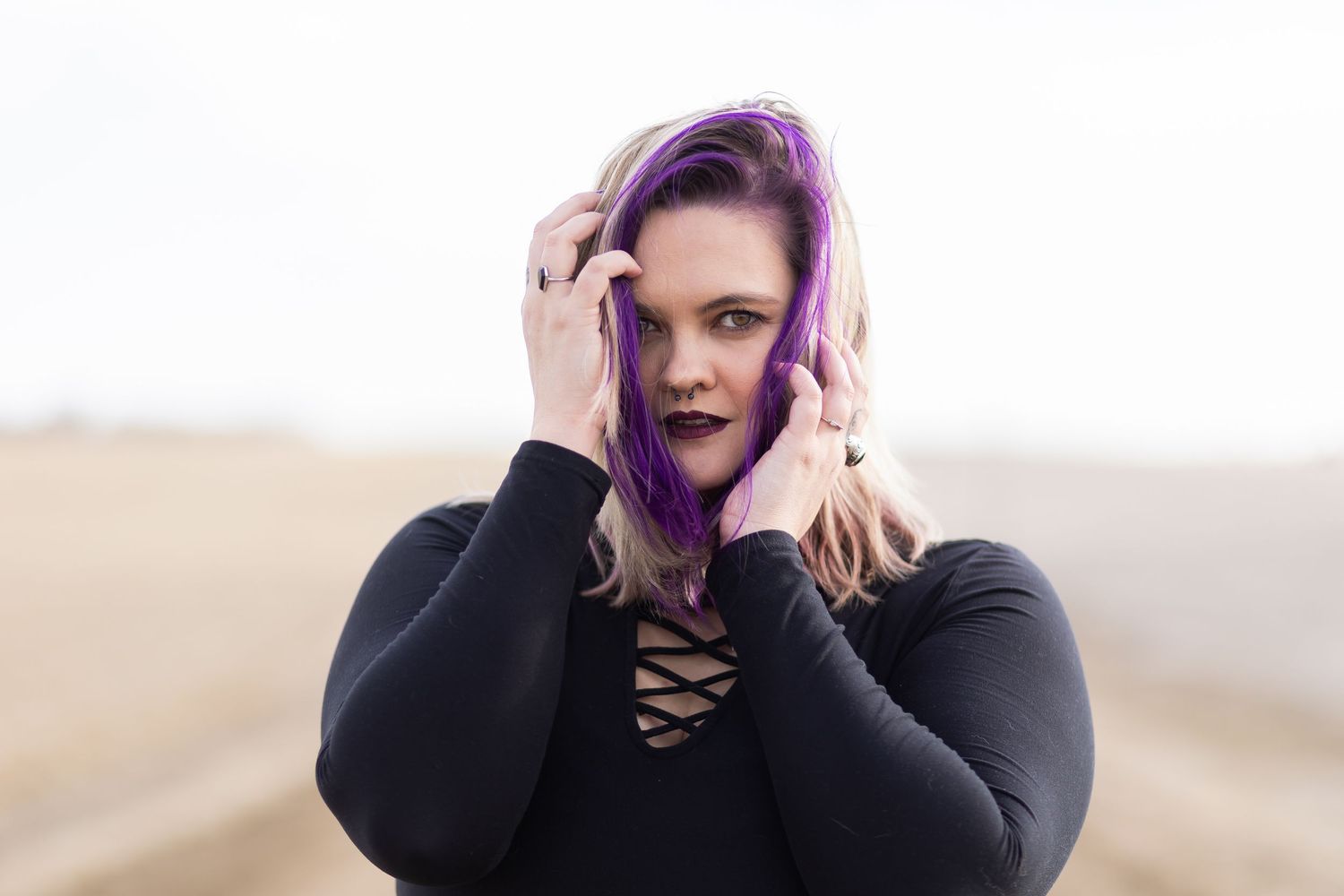 Woman with vibrant purple hair wearing a black hoodie poses against a soft beige background outdoors.