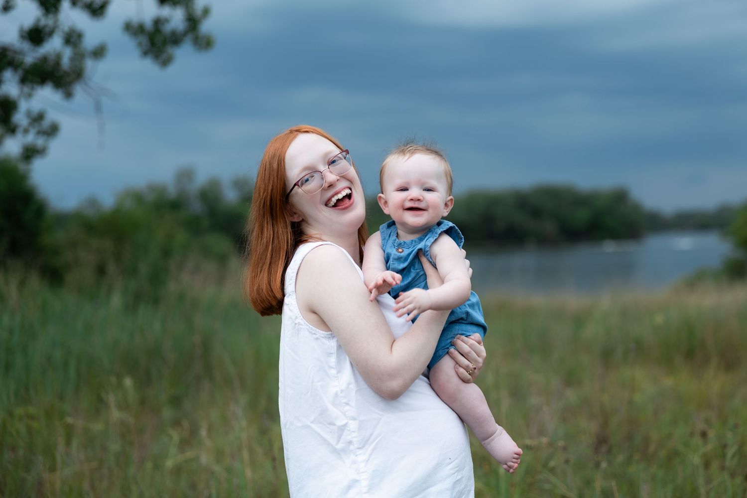 A joyful redhead woman in a white dress holds a smiling baby in denim outdoors near a lake.