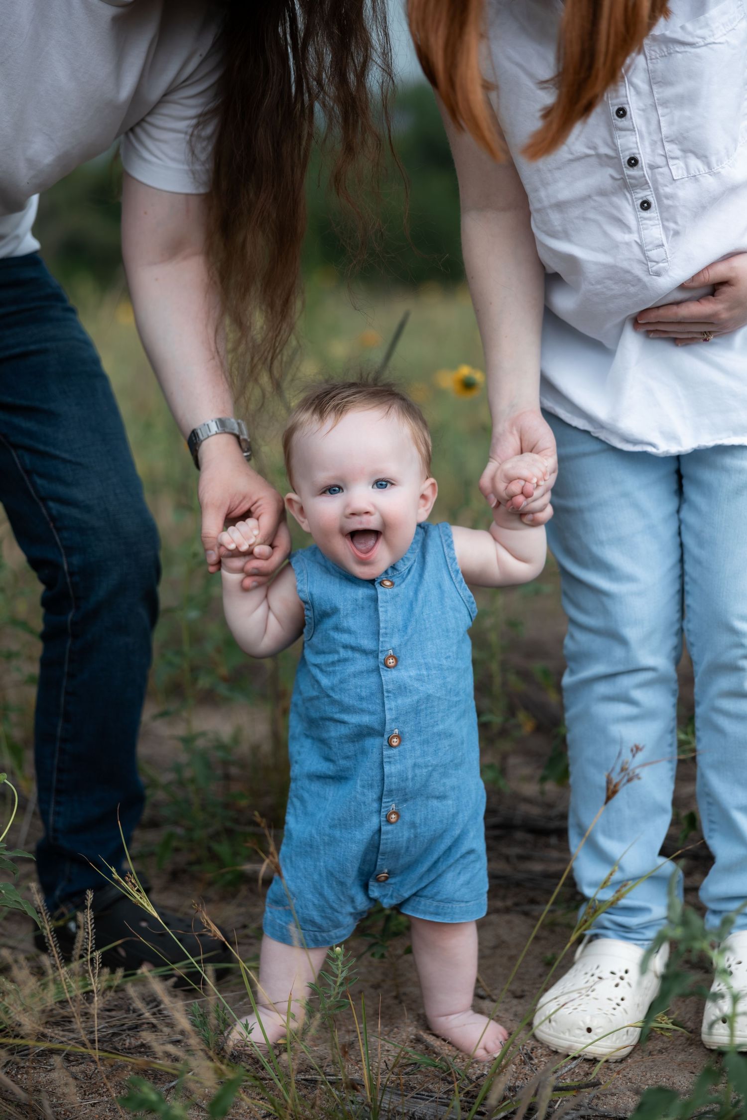 A joyful baby in a blue romper takes first steps outdoors, held by two adults in a sunny field.