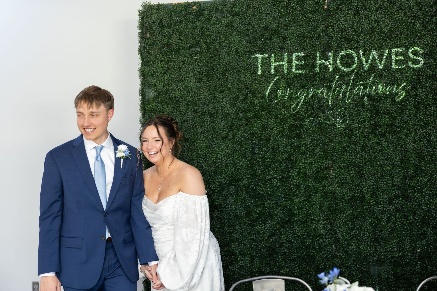 Newlyweds smile in front of a green hedge wall with The Howes Congratulations sign at their wedding reception.