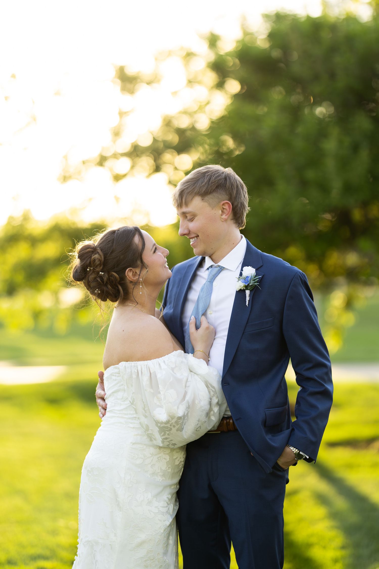 A bride and groom share a romantic moment outdoors, smiling at each other in golden sunset light.