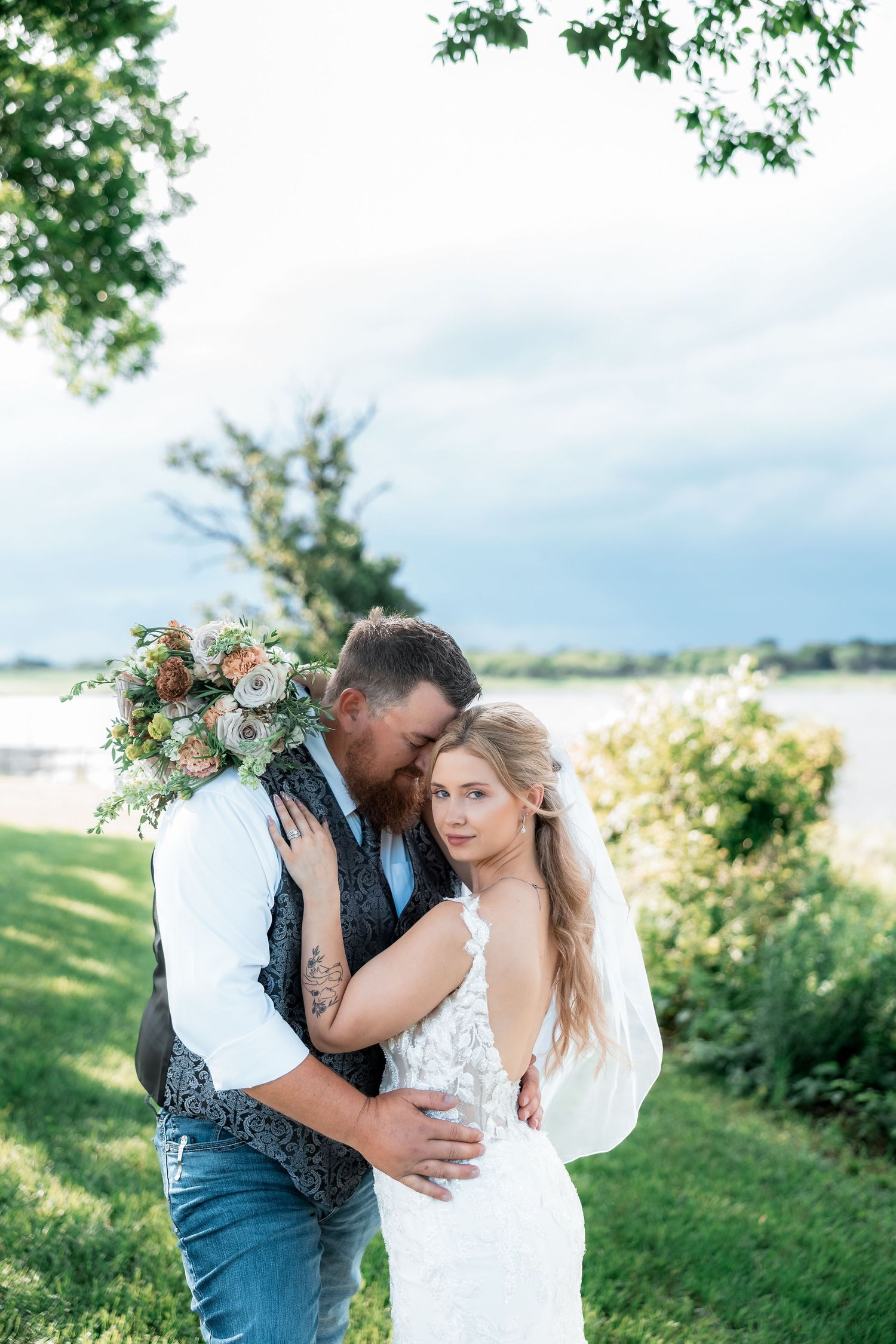 Bride and groom embrace outdoors near a lake, she holds a floral bouquet in a lace gown as he kisses her cheek.