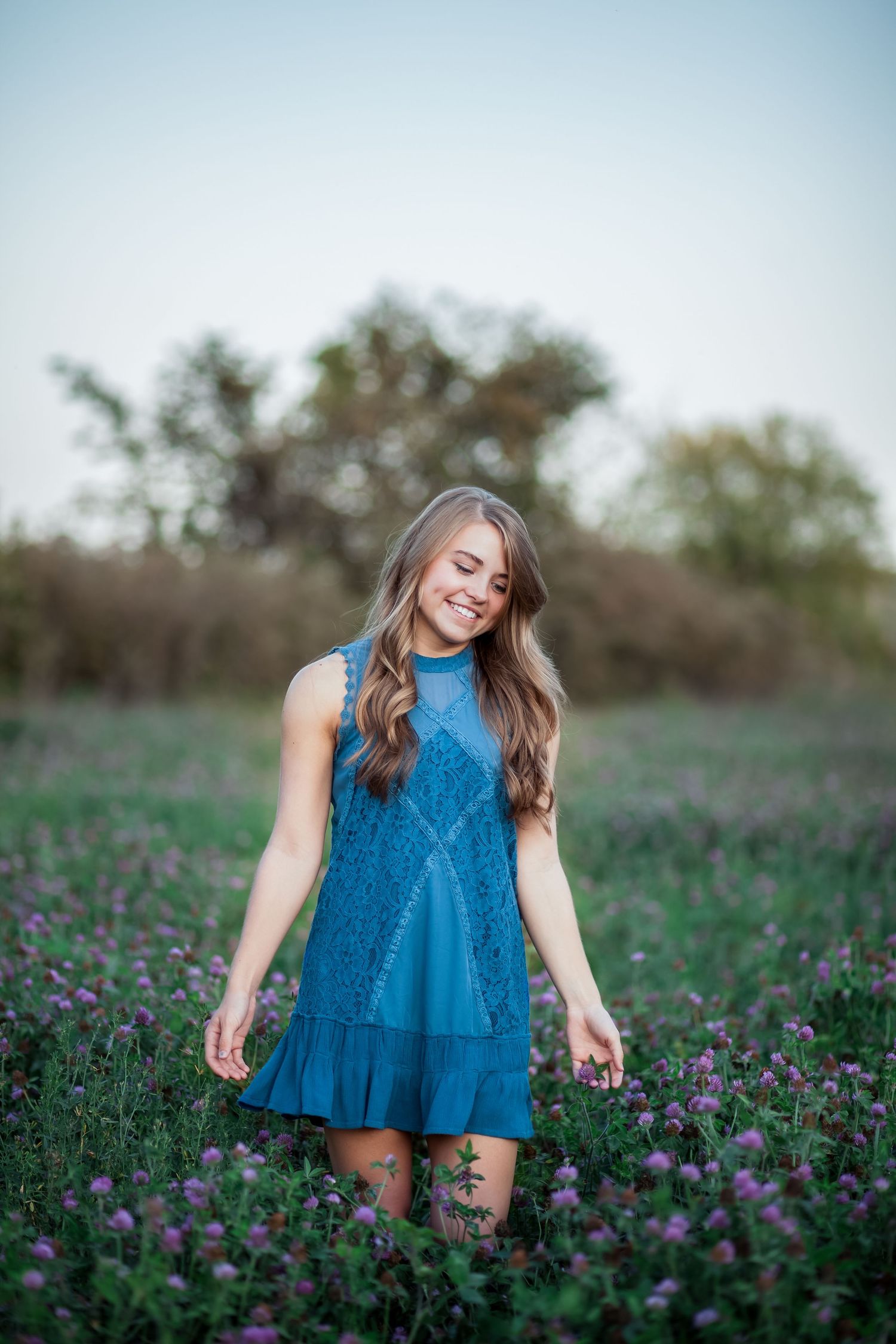 Young woman in blue lace dress smiling in a purple wildflower field at golden hour.