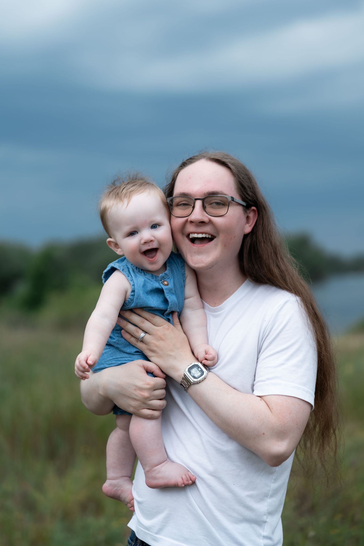 A laughing young woman with glasses holds a happy baby outdoors against a cloudy sky backdrop.