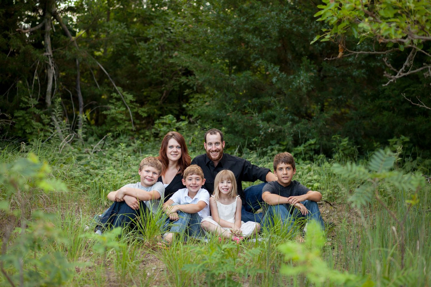 A group sits together in a lush green field surrounded by trees and summer foliage.