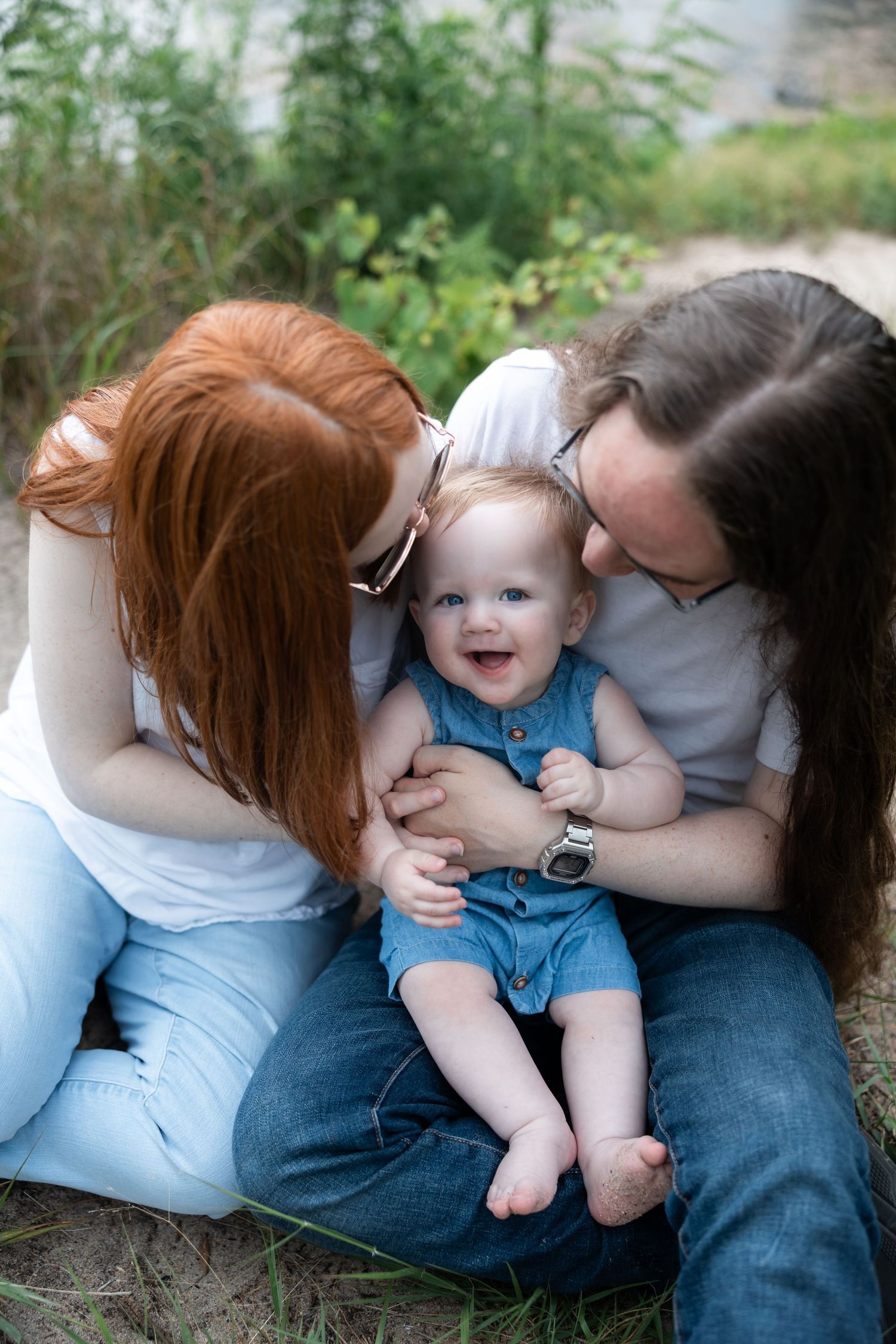 Two parents kissing their happy smiling baby outdoors, sitting together on grass with greenery in the background.