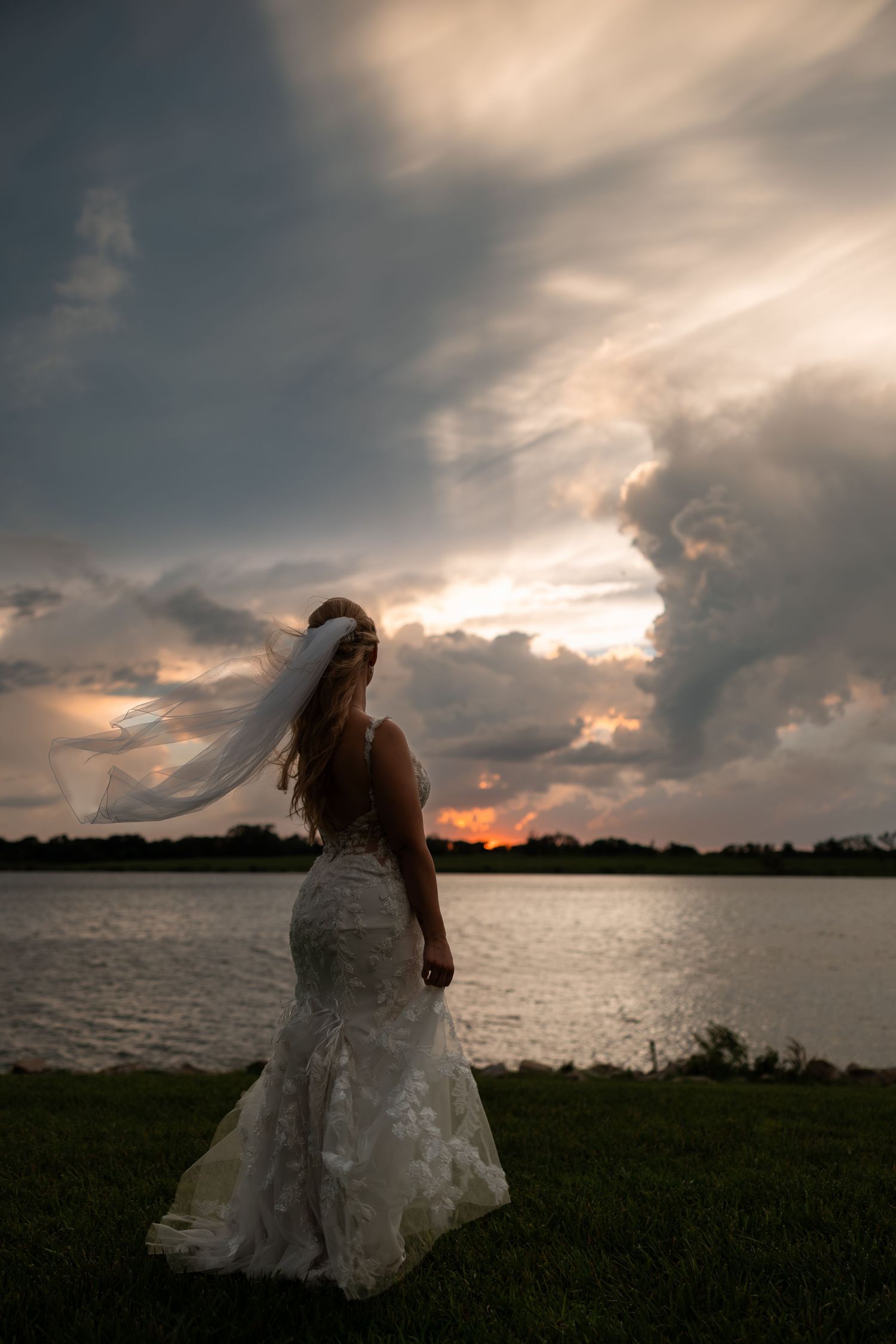 Bride in lace mermaid gown and veil stands by lakeside at dramatic sunset with stormy clouds.