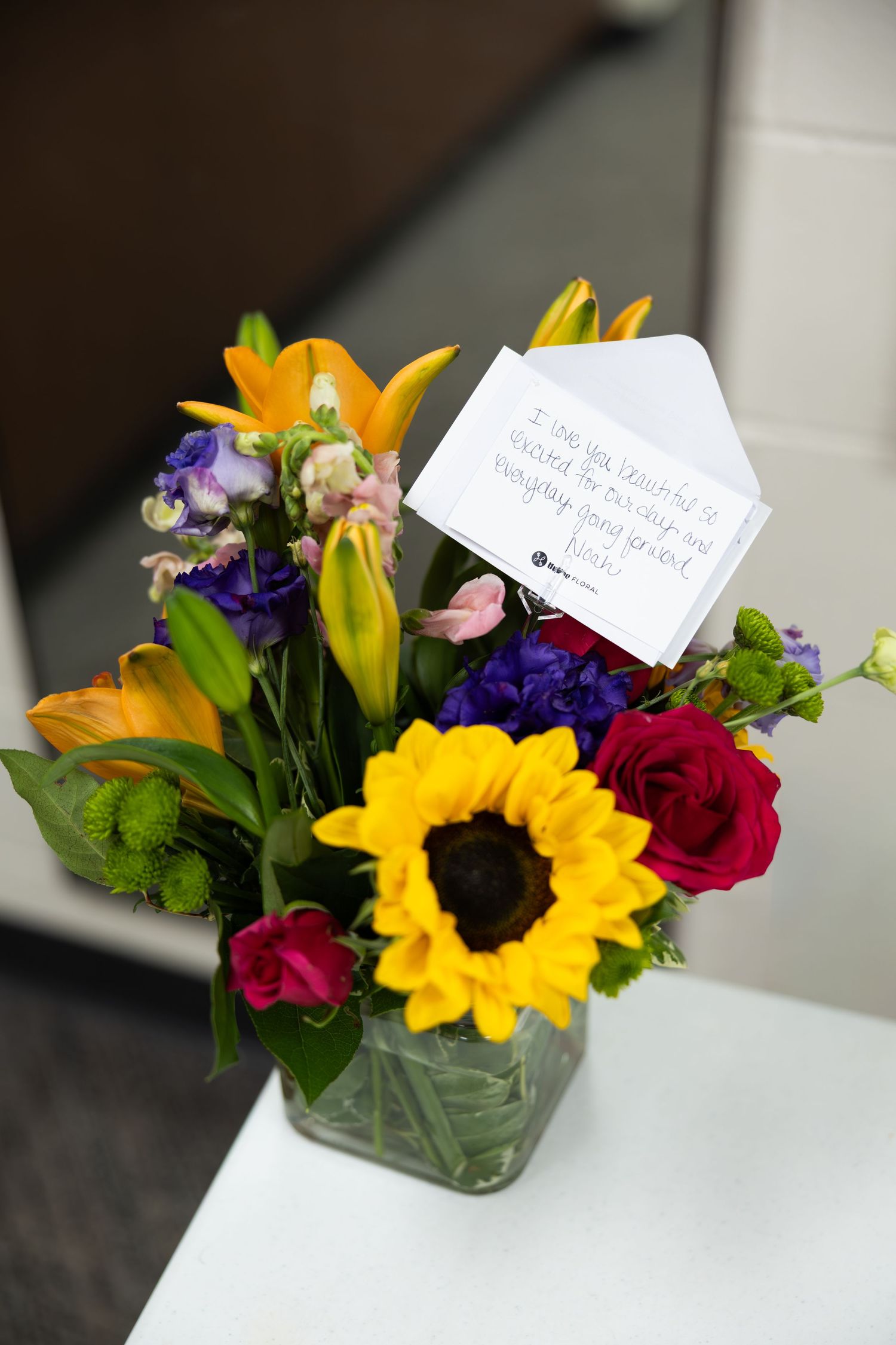Colorful flower bouquet with sunflower, roses, lilies, and a handwritten note card in a glass vase on a white table.