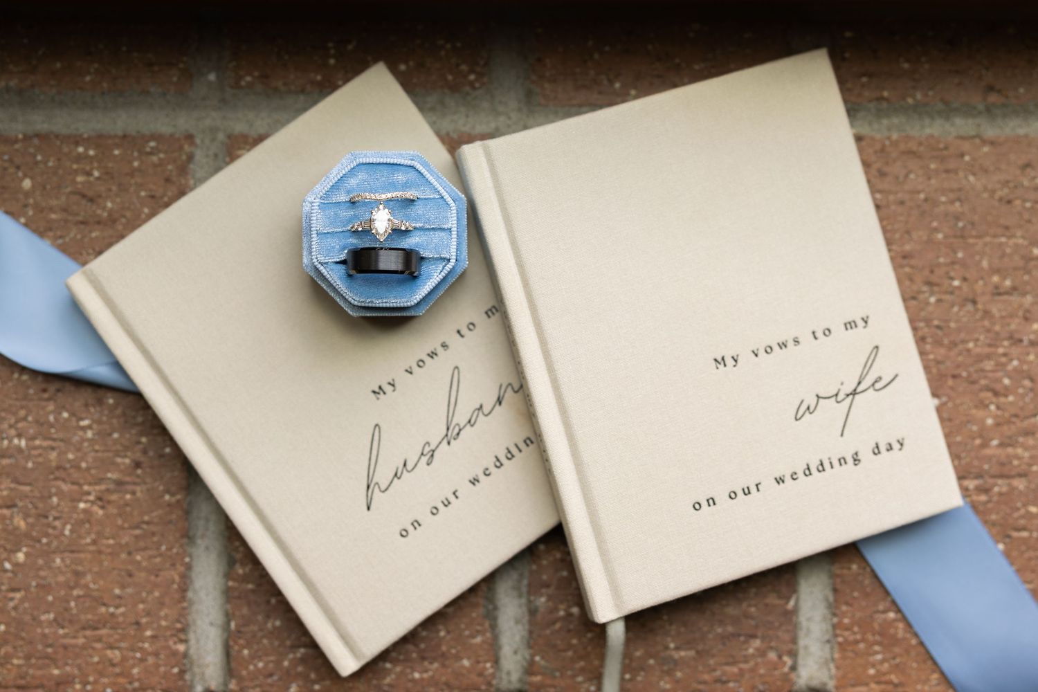 Wedding vow books for husband and wife with a blue velvet ring box containing engagement rings on a brick surface.