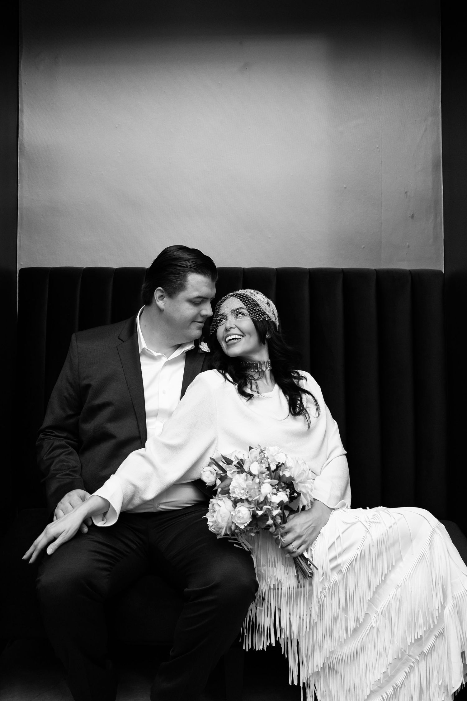 A vintage-inspired black and white wedding portrait of a couple sitting together with a floral bouquet.