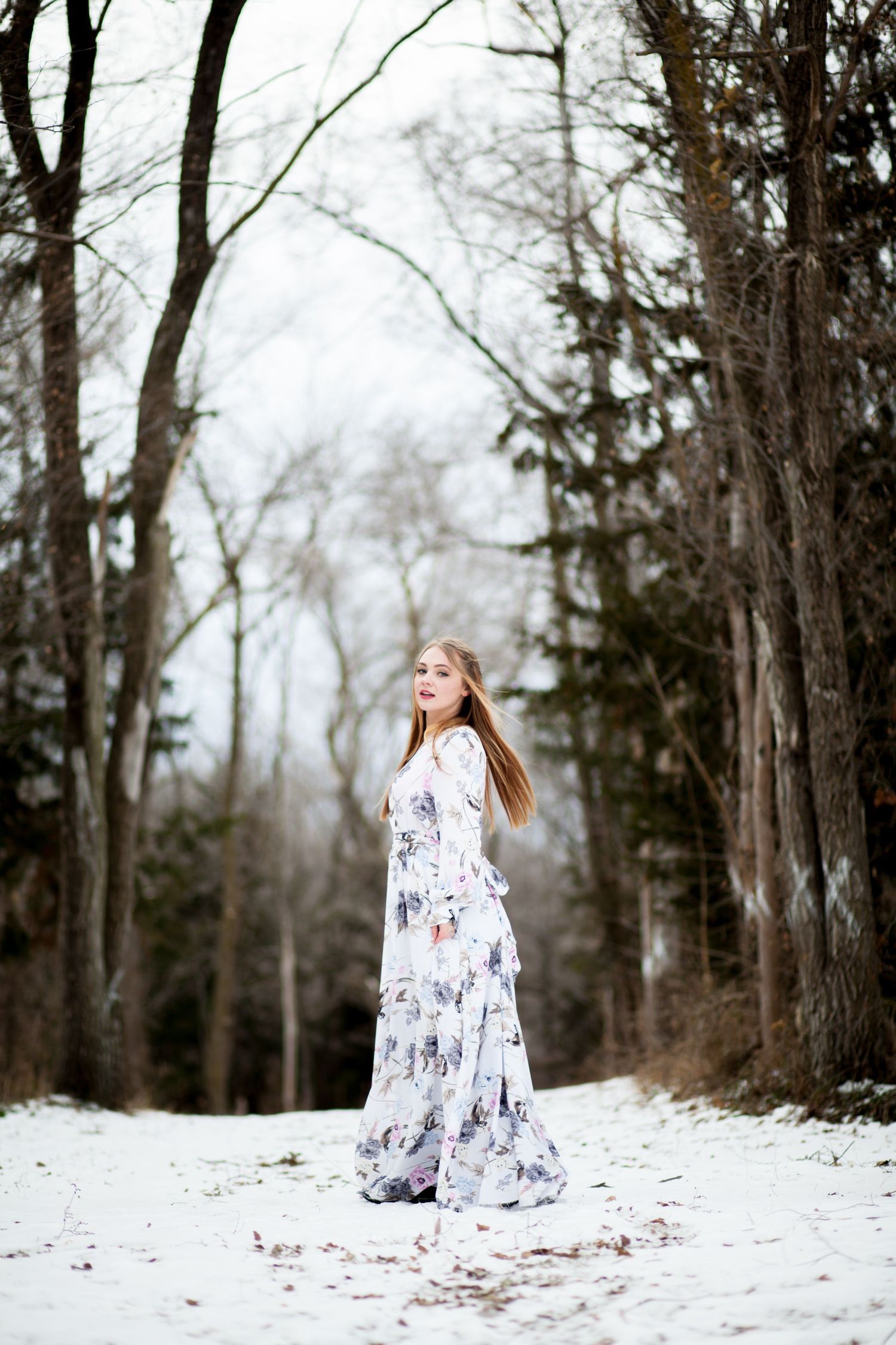 A white floral dress contrasts against snowy woods in an ethereal winter portrait.
