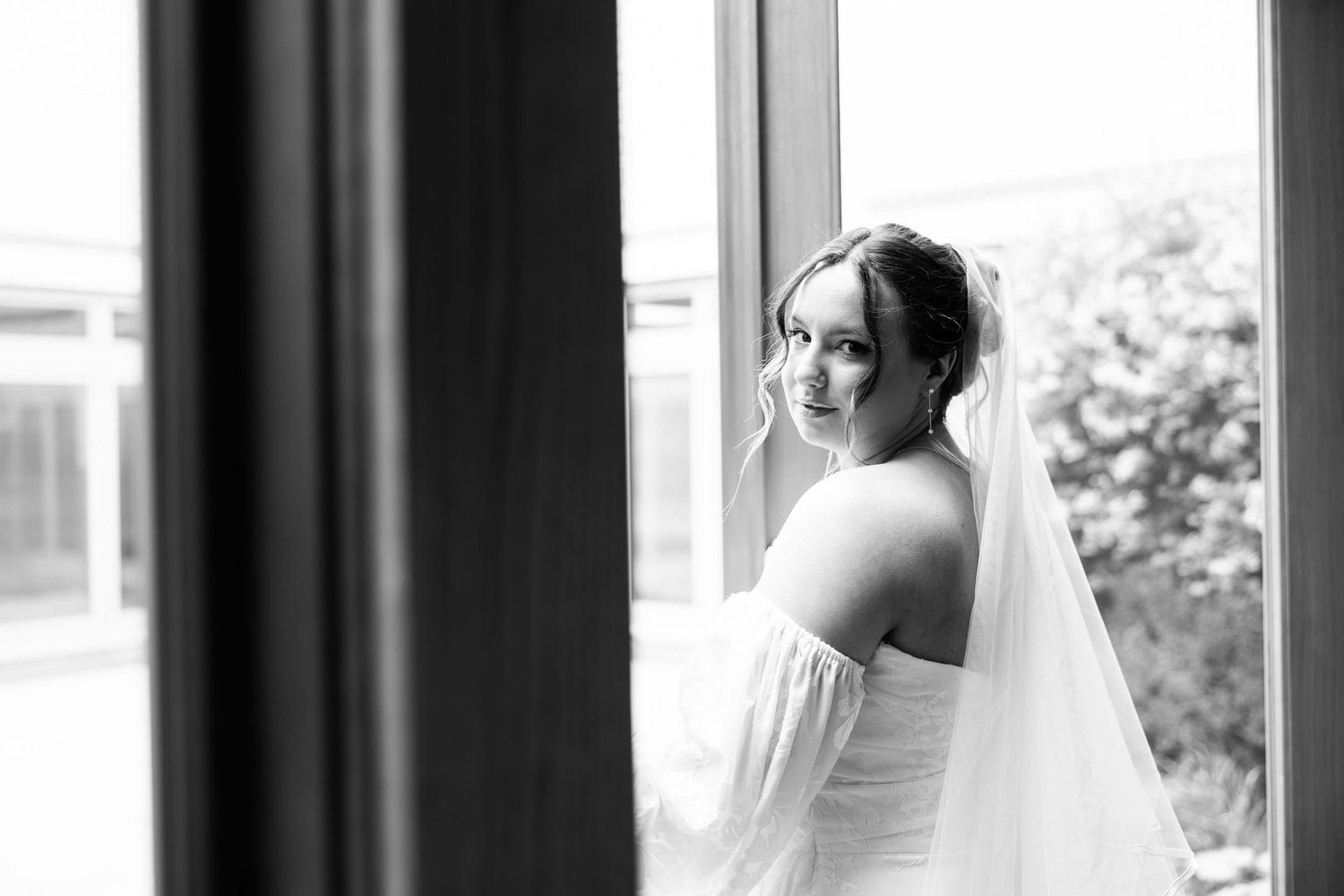 Black and white photo of a bride in a white off-shoulder gown and veil standing by a window.