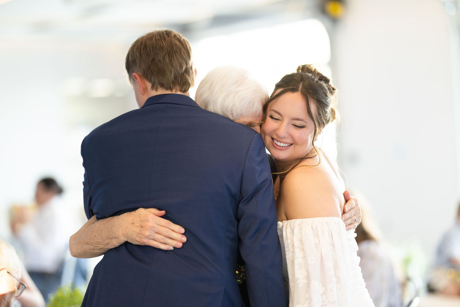 Bride in white dress smiling while sharing a warm group hug at a wedding reception.